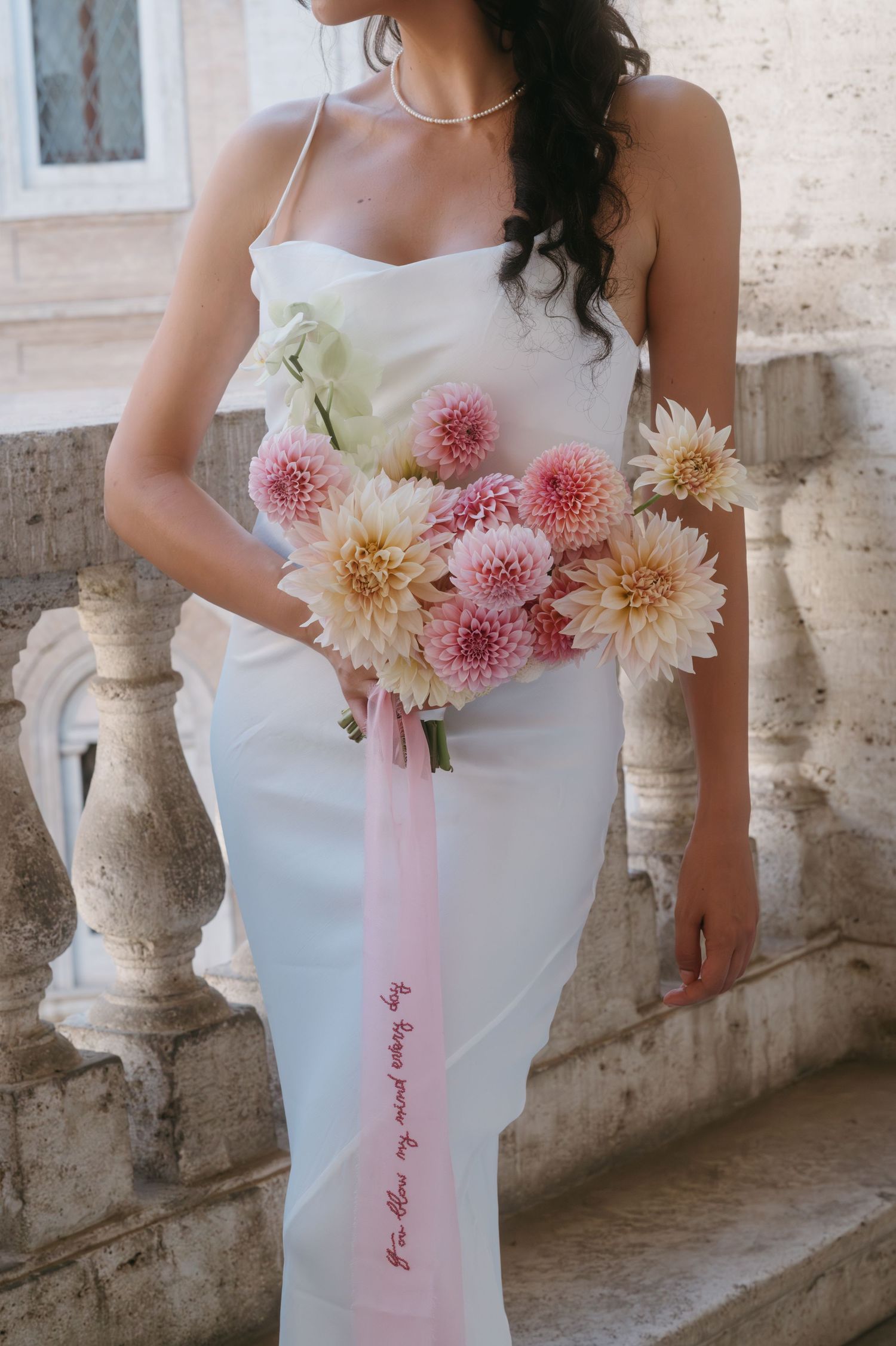 Elegant white wedding dress with pink and cream dahlia bouquet against stone architecture.