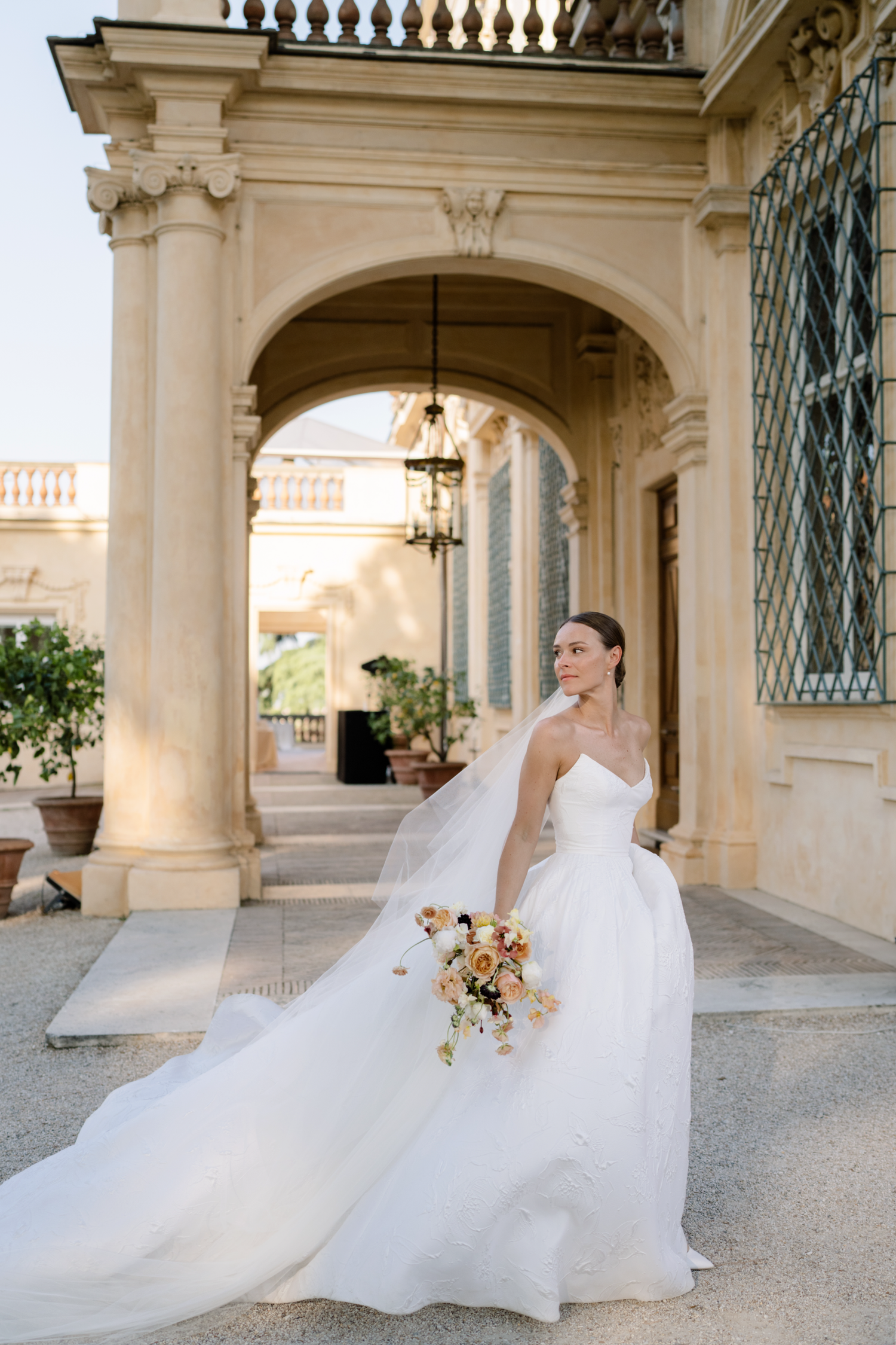 Elegant bride in flowing white gown with long train twirls gracefully under ornate stone archways.