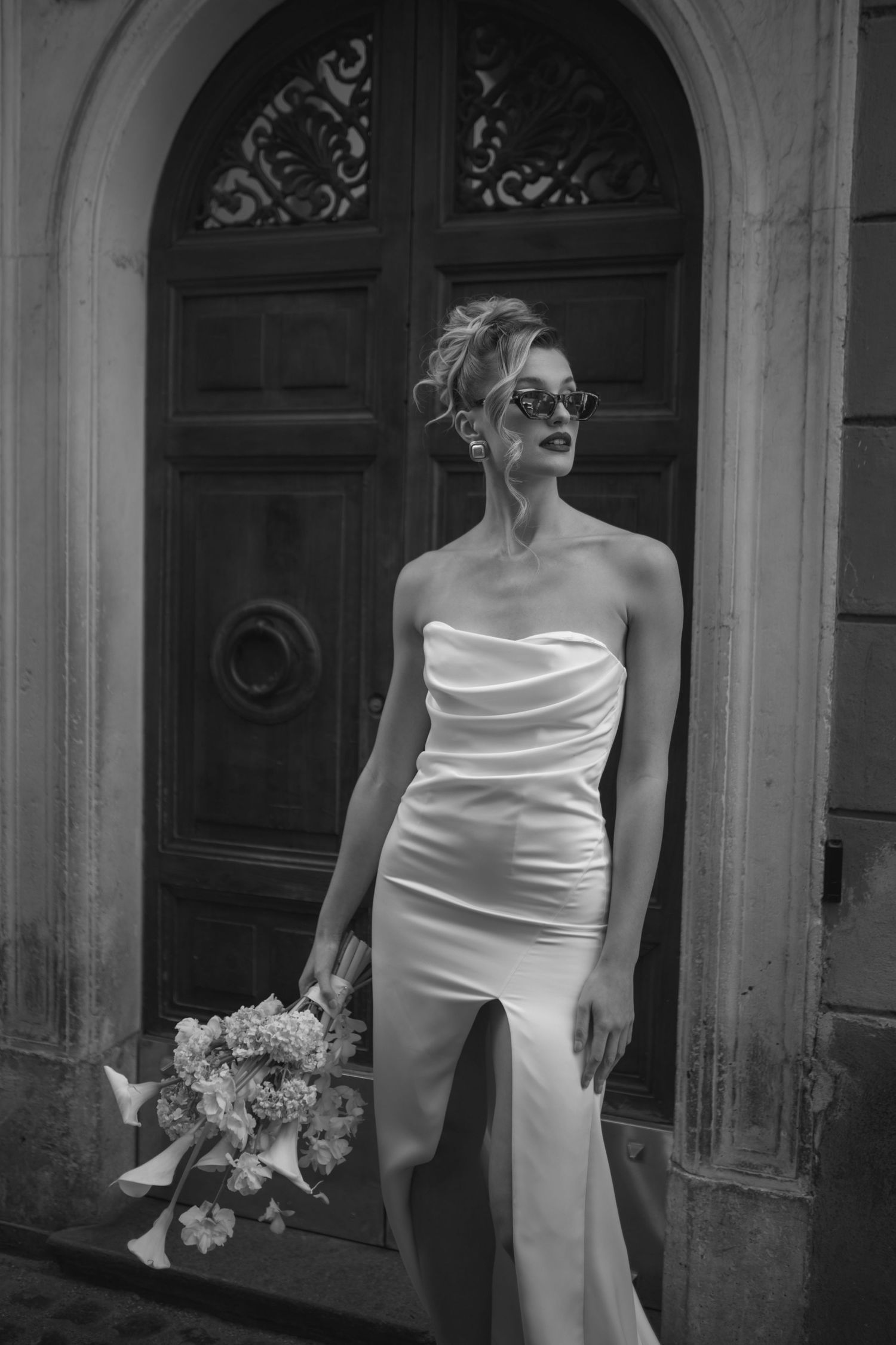 Elegant bride in a white off-shoulder dress poses against a classic architectural backdrop.
