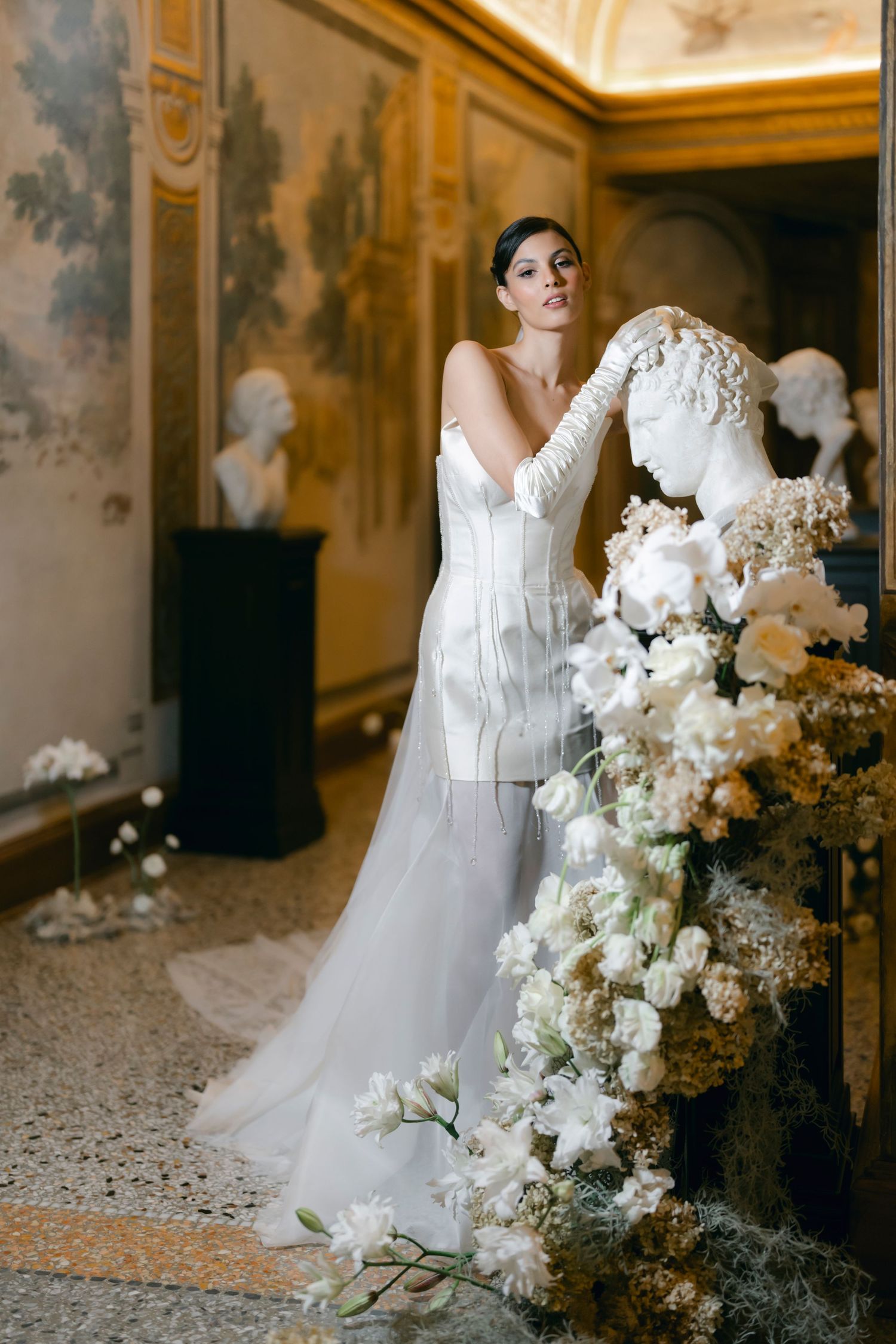 Elegant white wedding dress with lace details and cascading floral arrangements in an ornate interior setting.
