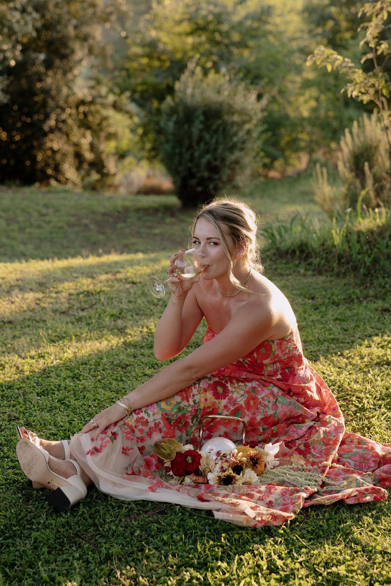 Woman in red floral dress sits on grass with wine glass during golden hour photography session.