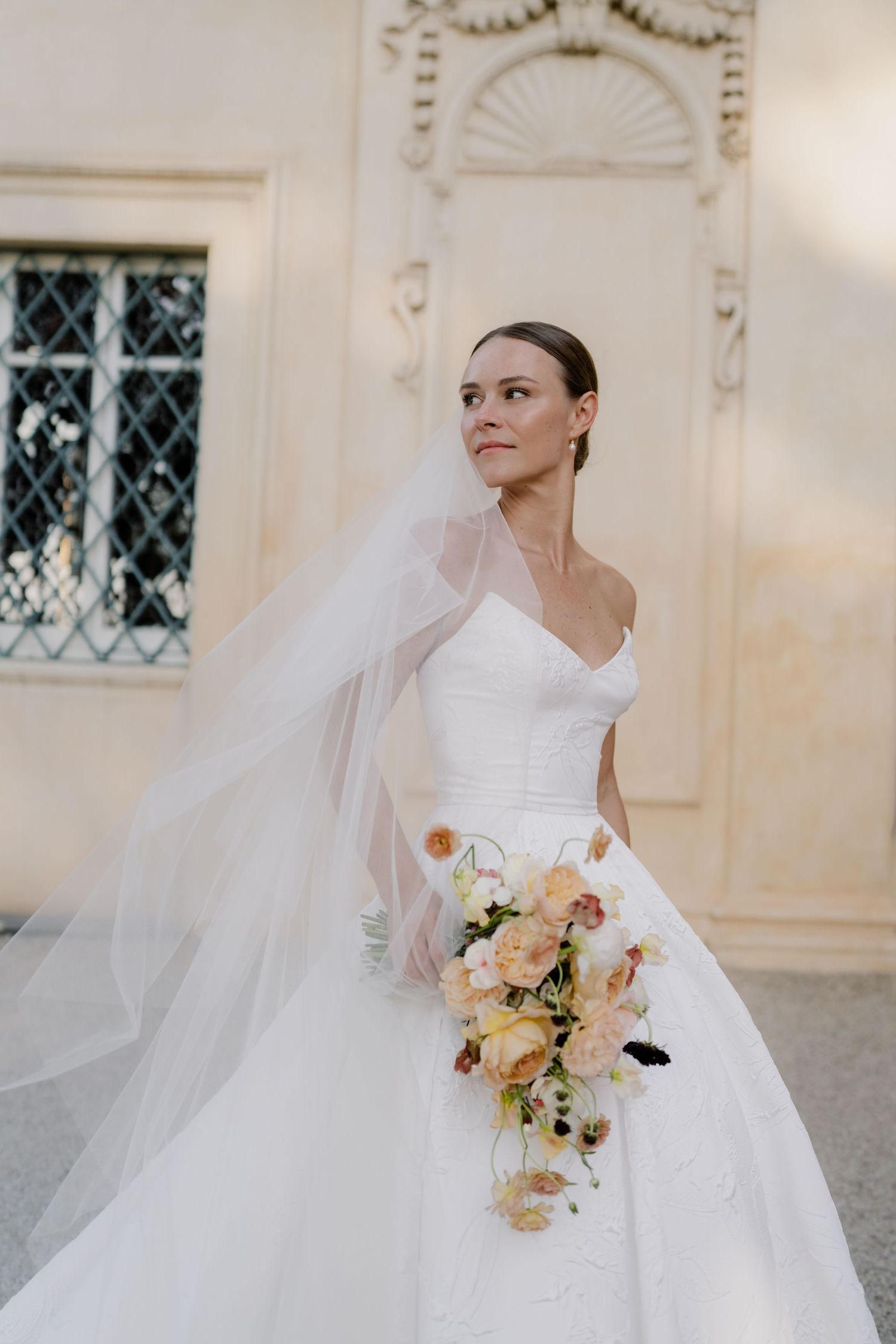 Elegant bride in white ball gown and veil holding peach and cream floral bouquet in ornate venue.