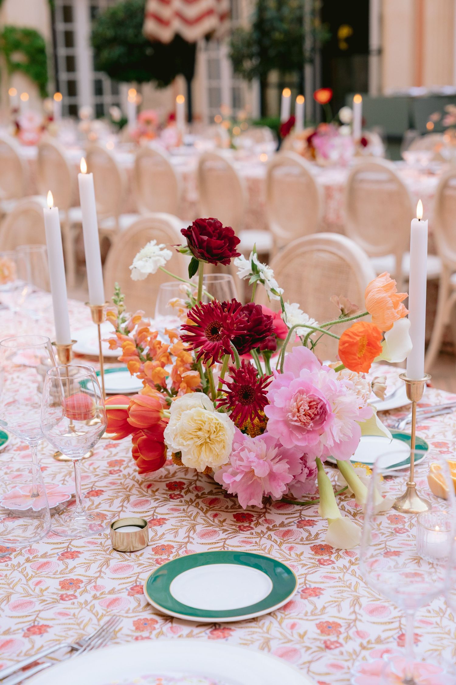 Romantic table setting with deep red and pink flower arrangement, white candles, and patterned tablecloth.