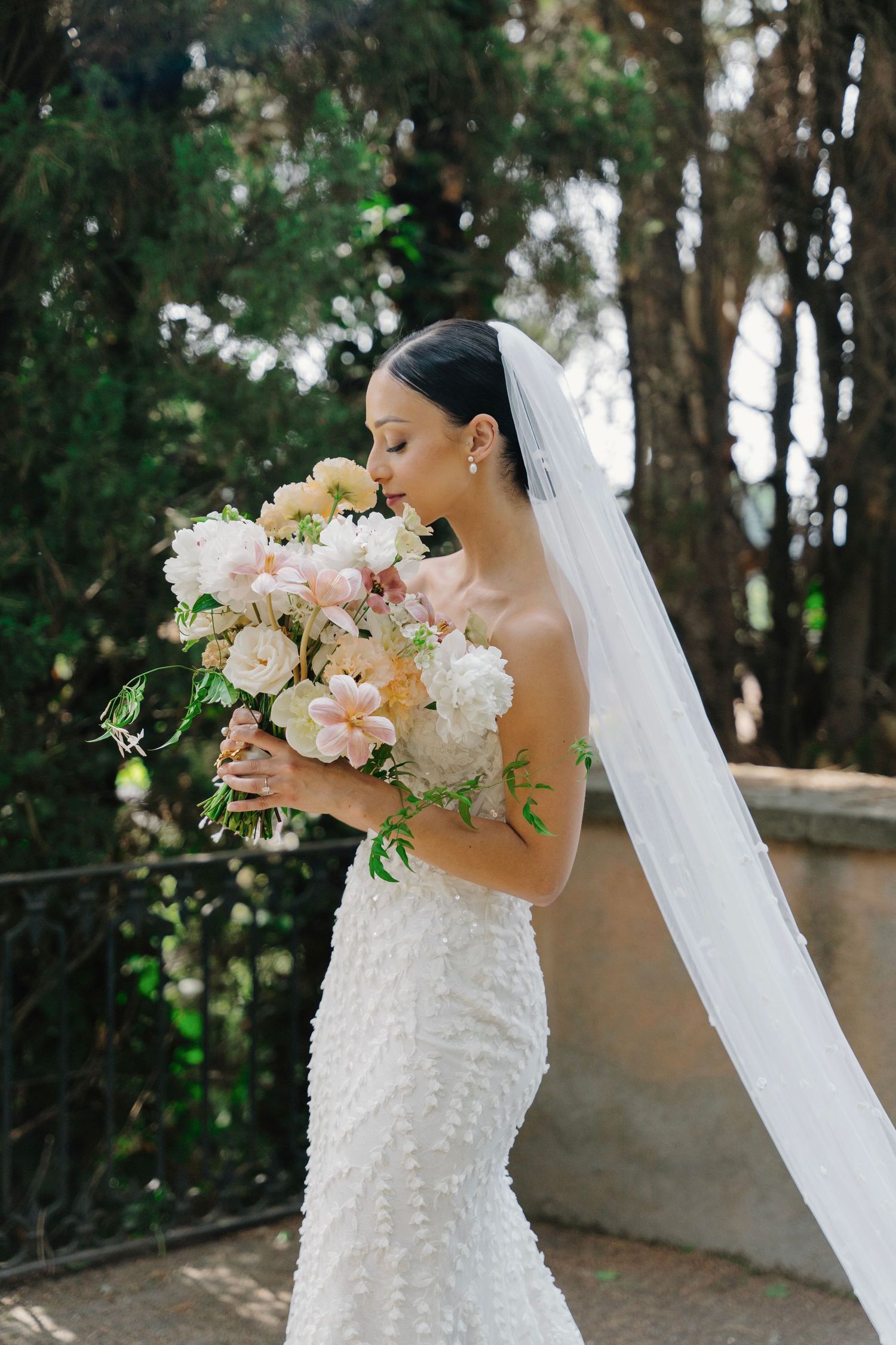 White textured wedding gown with flowing veil and soft pink floral bouquet in garden setting.