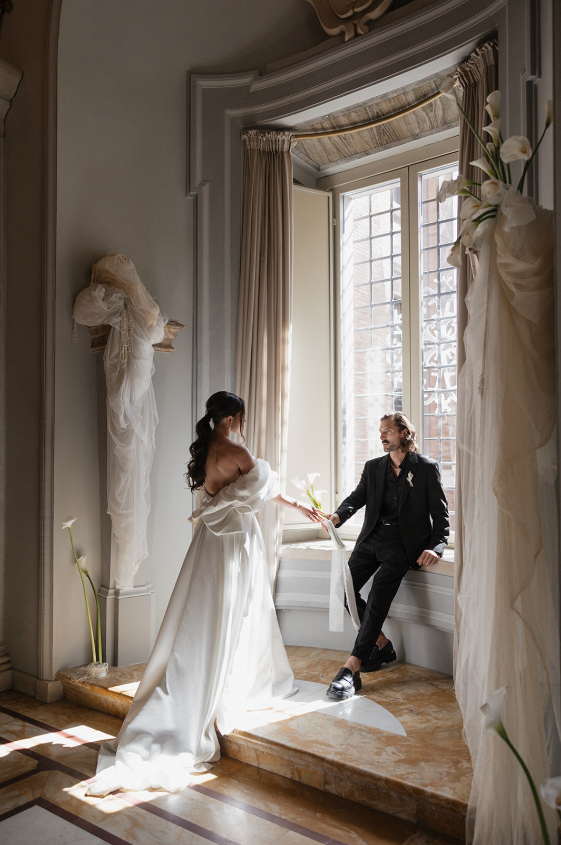 Sunlit wedding portrait in elegant room with large windows and flowing white dress.
