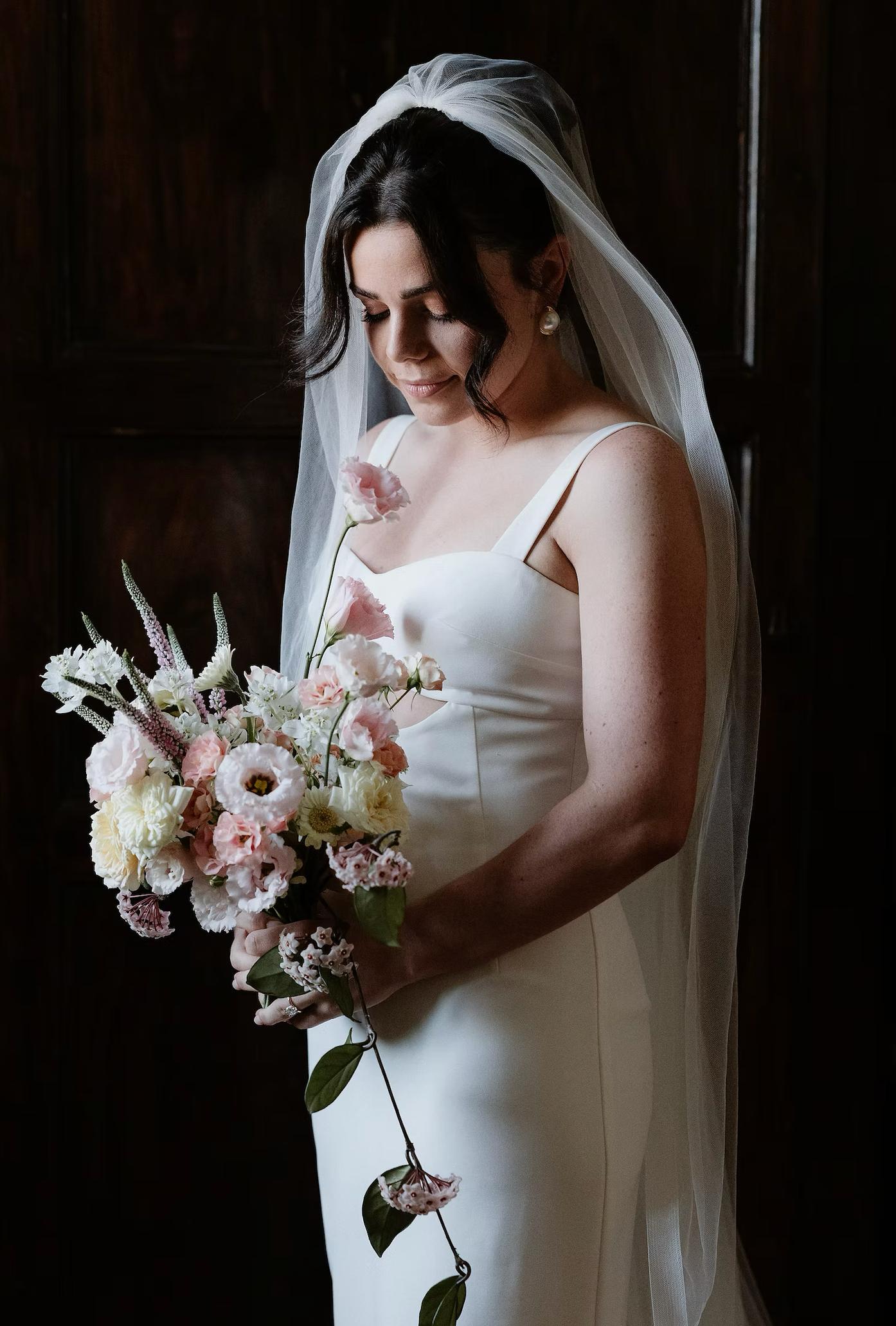 Bride in sleek white gown holding a romantic bouquet of pink and white flowers against dark background.
