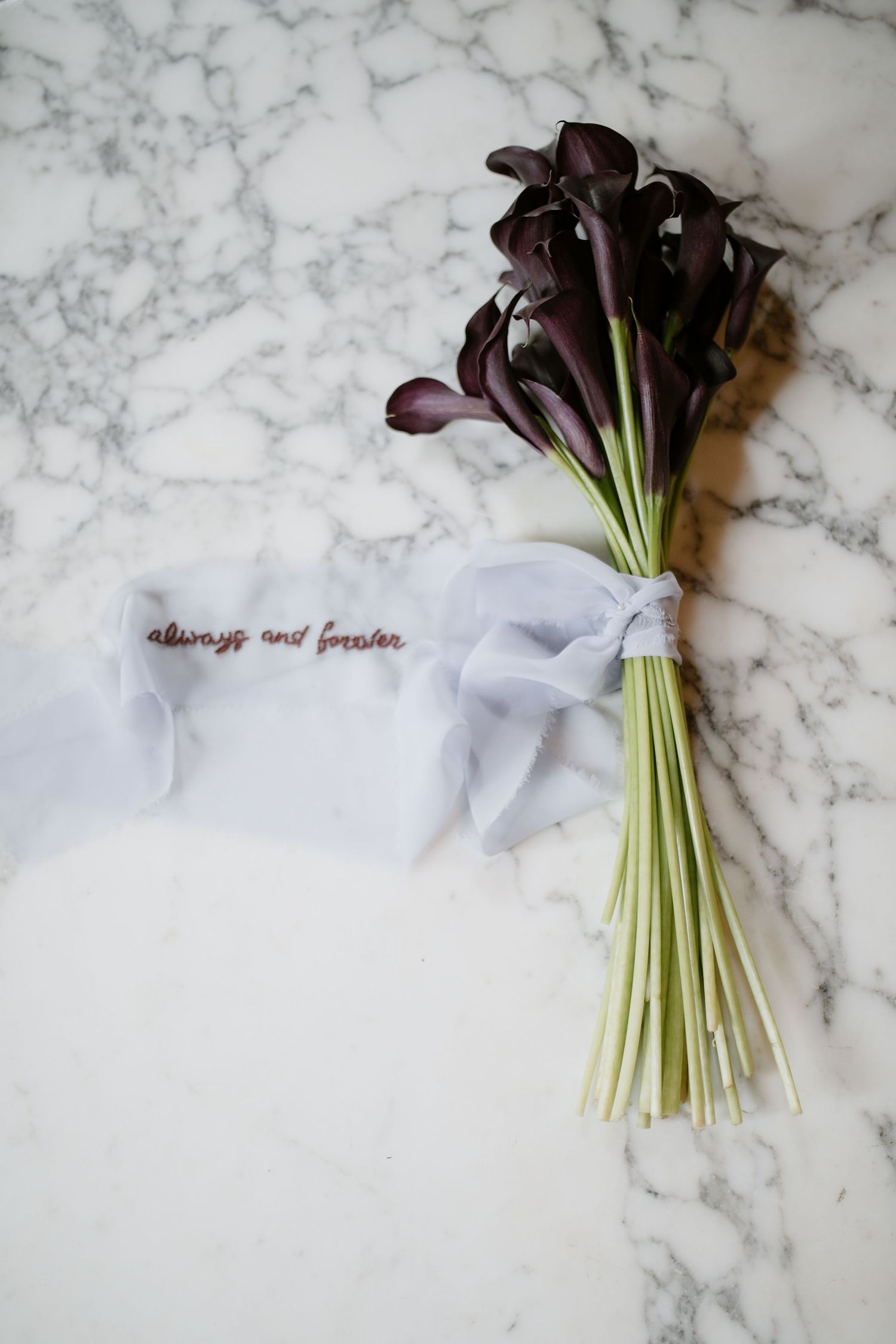 Deep purple calla lilies tied with white ribbon against marble background with embroidered text.