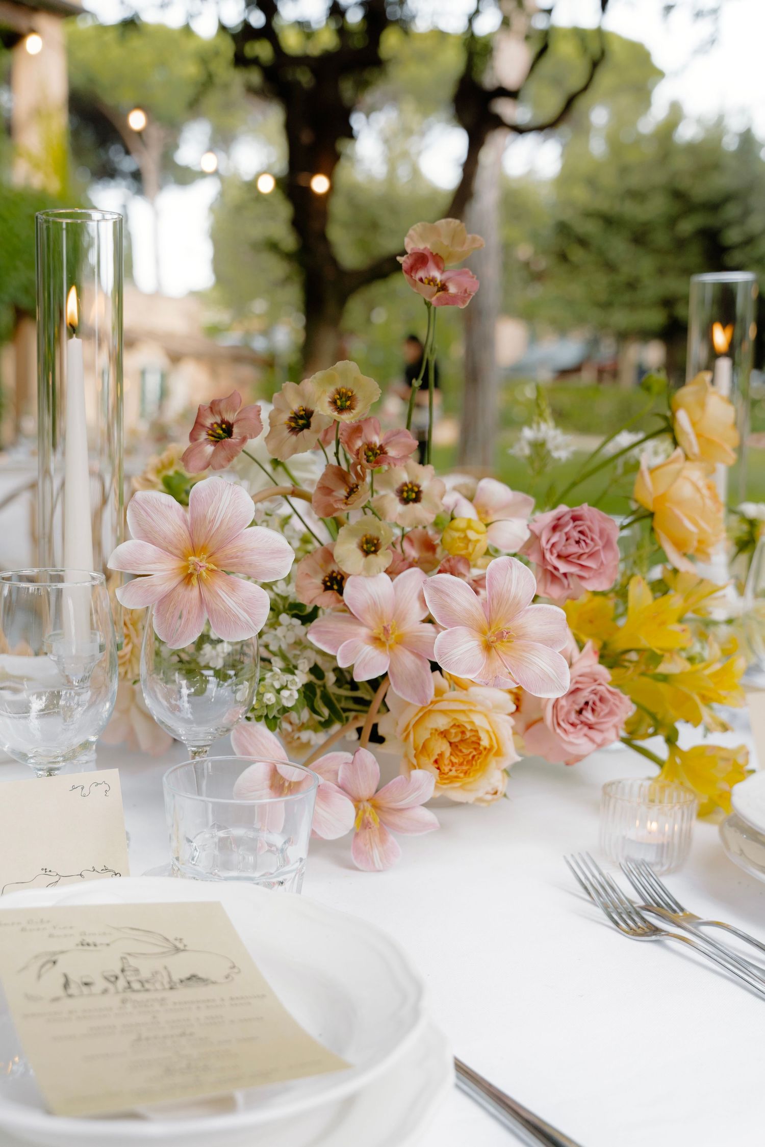 Elegant table setting with pink and yellow flowers in delicate glass vases for a romantic outdoor wedding reception.