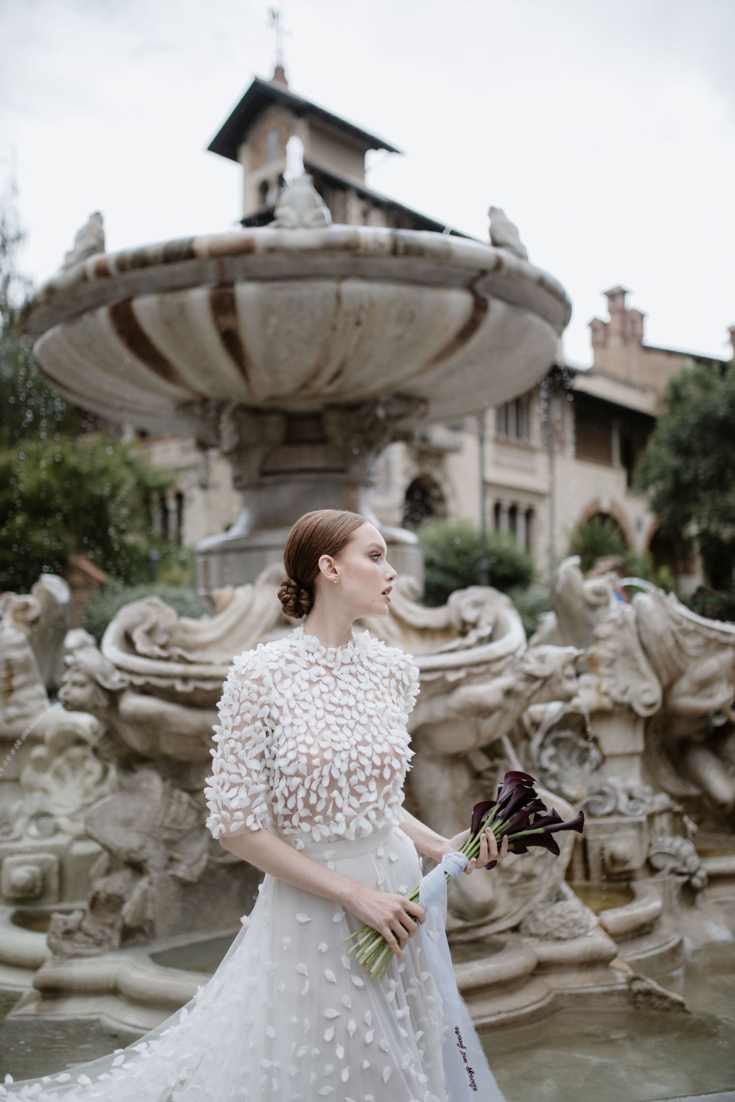 A bride holding dark flowers poses by an ornate stone fountain with architectural details.