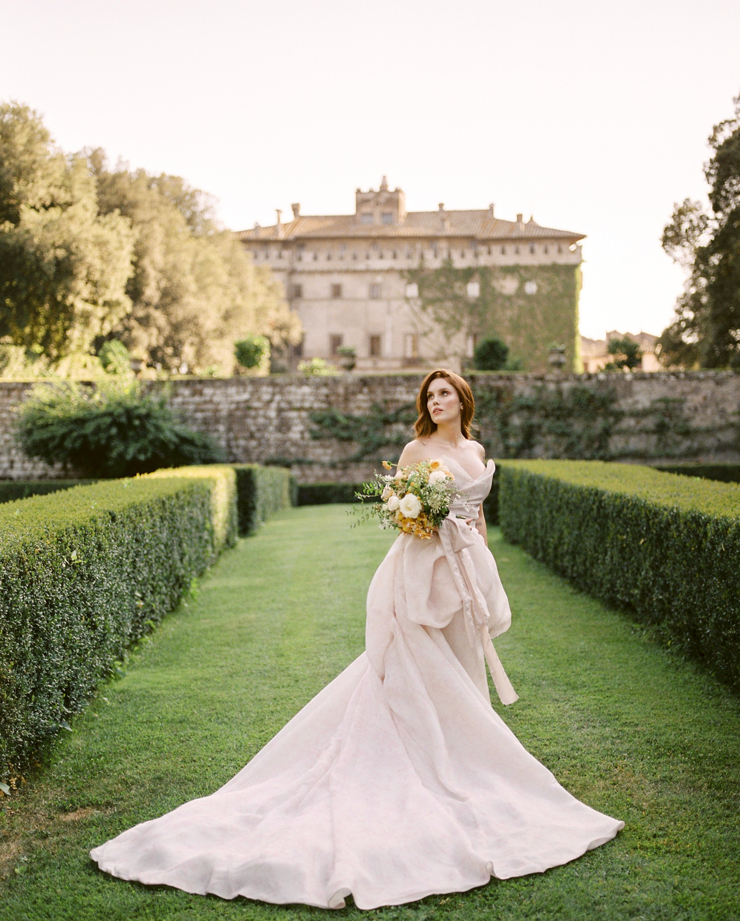A flowing white wedding gown trails elegantly through a manicured garden path with stone walls and hedges.