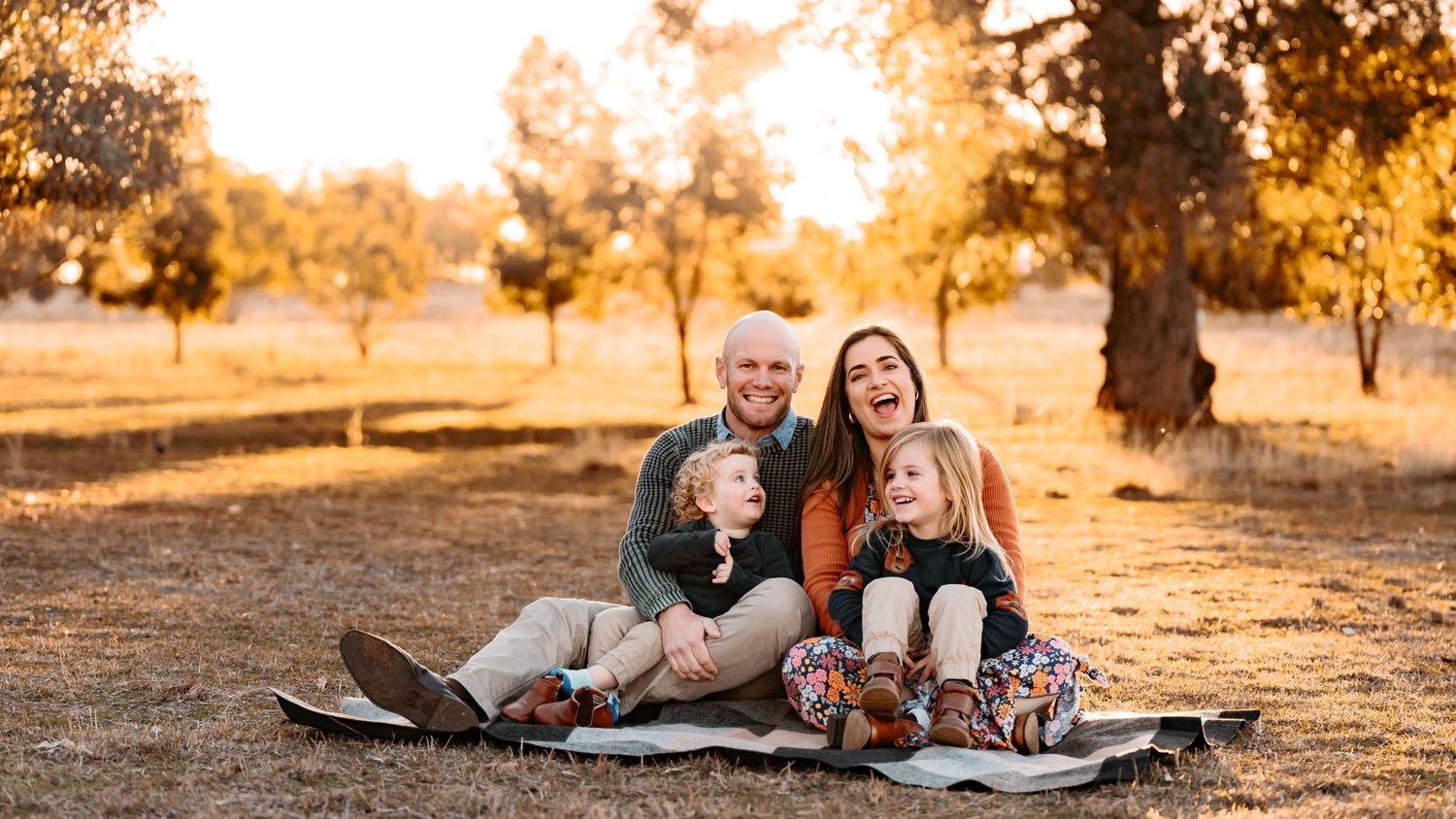 Pictured is a family of 4 - a mother, father & 2 young boys. They're having a candid moment on a mat. They're outdoors at sunset.