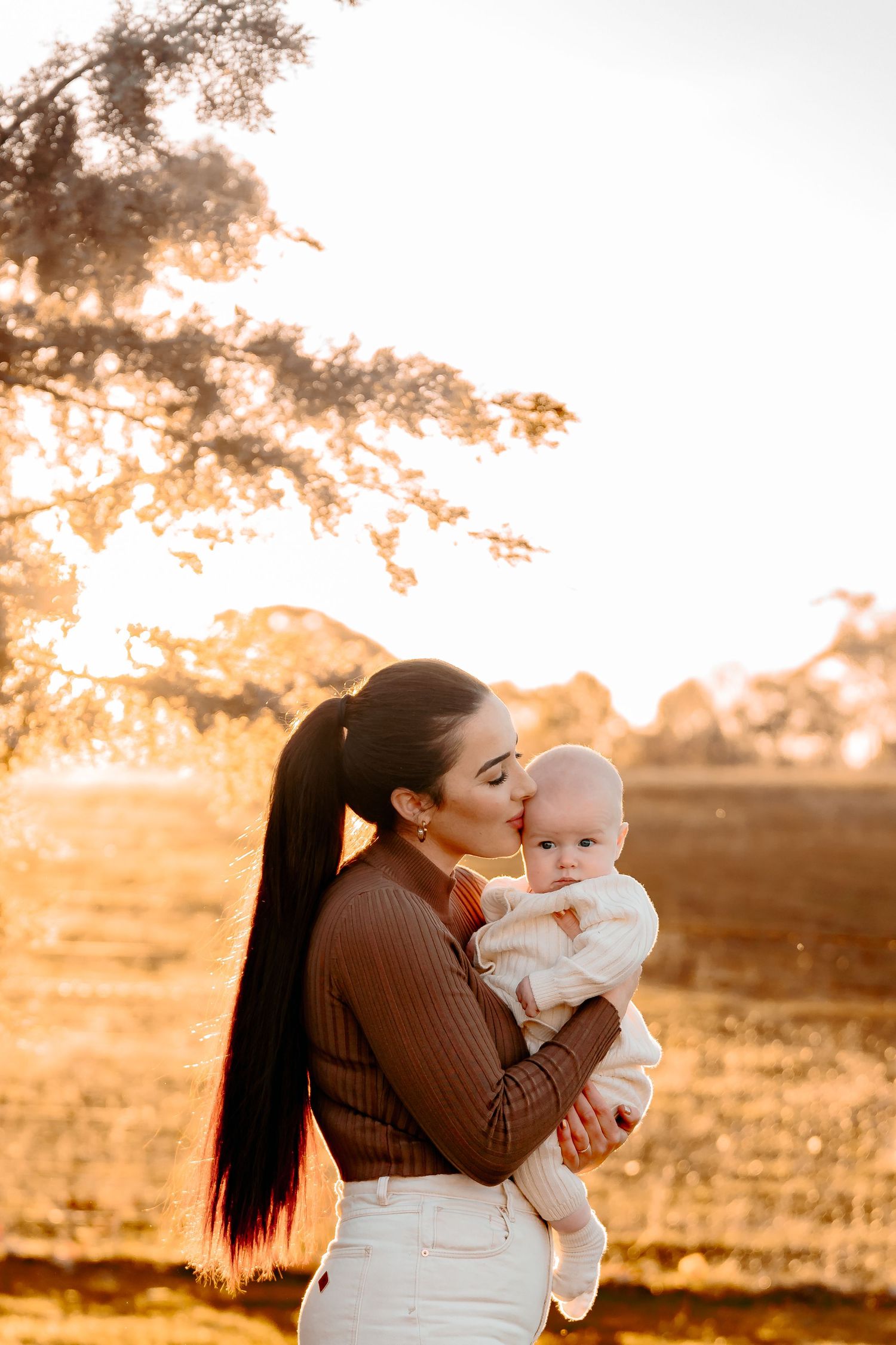 Stunning mother kissing her baby on the head with beautiful backlit light at sunset. Mother has lucious long hair & is doting on the child
