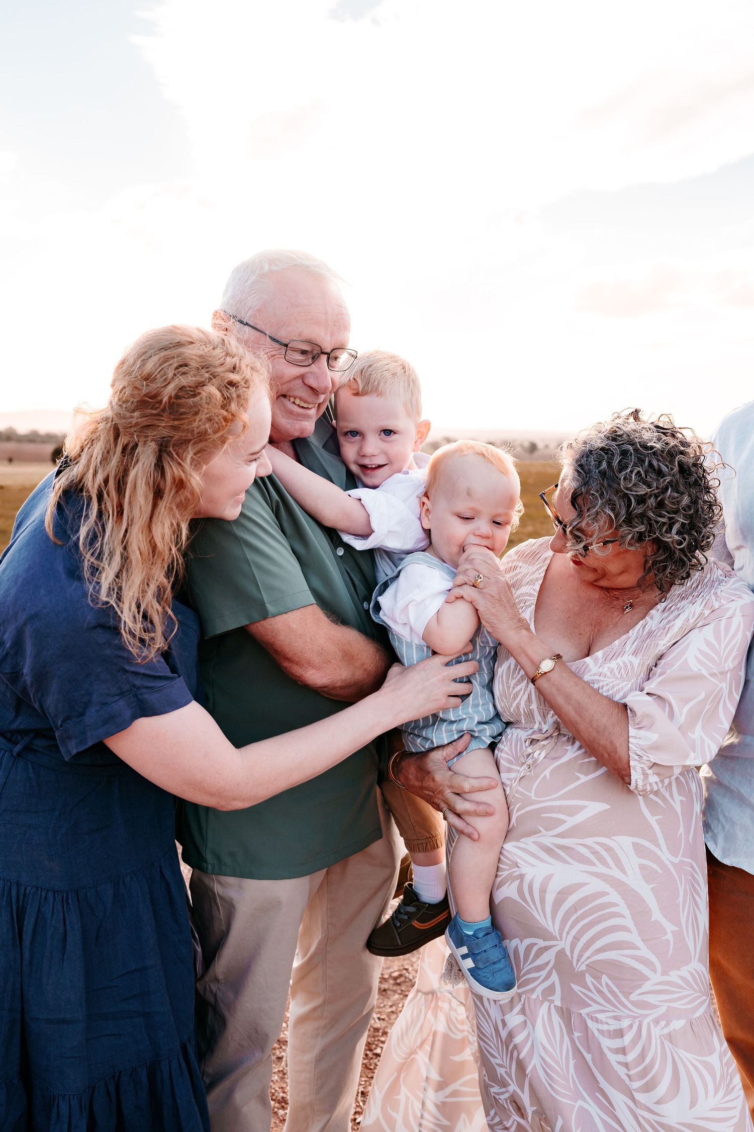 A MOTHER, HER PARENTS AND HER TWO LITTLE BOYS LAUGHING AT THE YOUNGEST CHILD WHO IS TRYING TO SHOVE HIS HAND IN HIS MOUTH