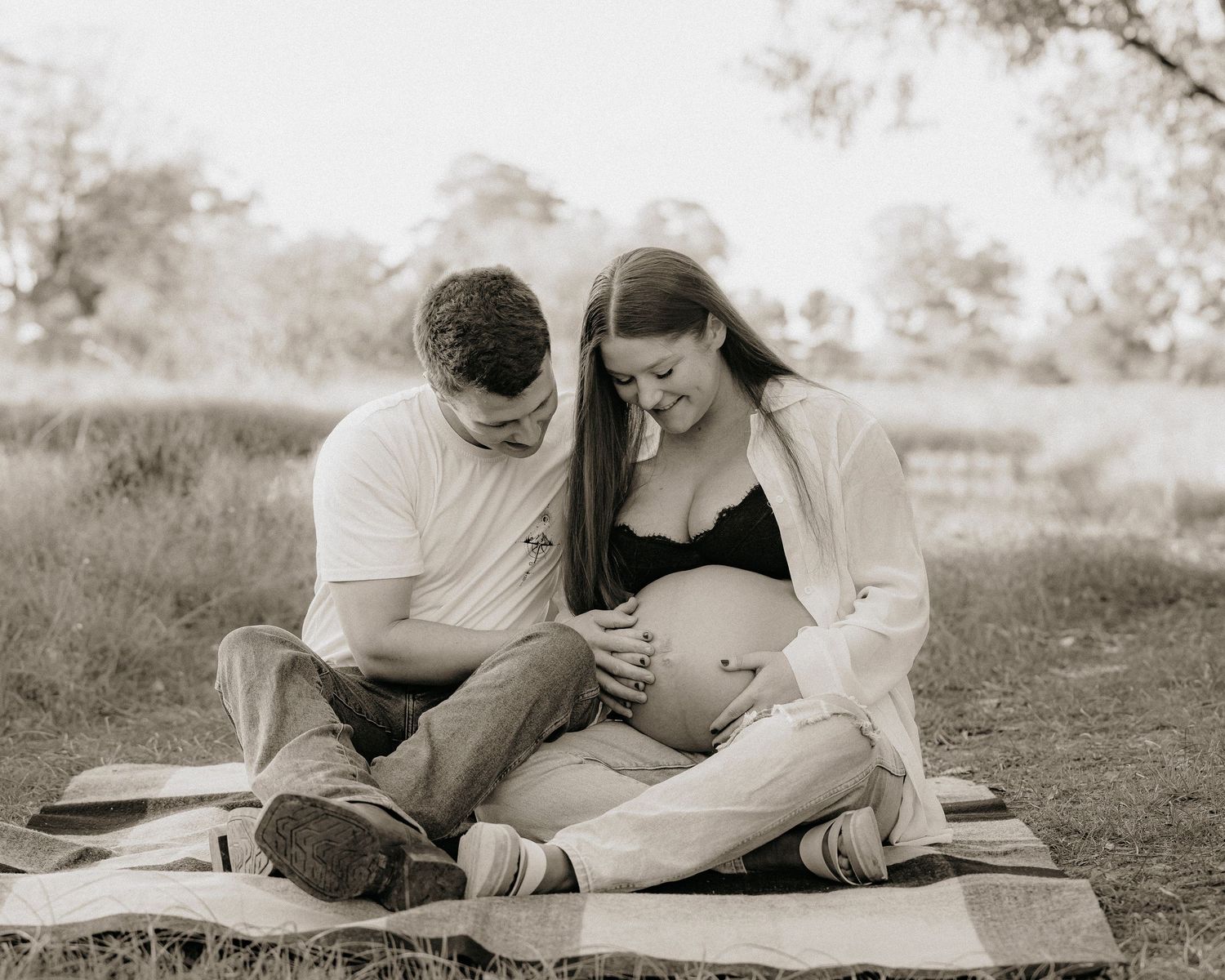 black and white image of proud parents to be sitting on a rug looking down at their baby growing inside mums glowing belly