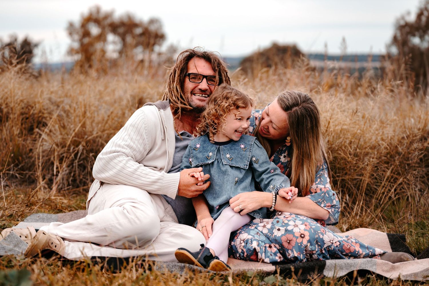 a dad and mum connecting with their daughter. they are all giggling and holding hands whilst enjoying a special moment seated on a mat