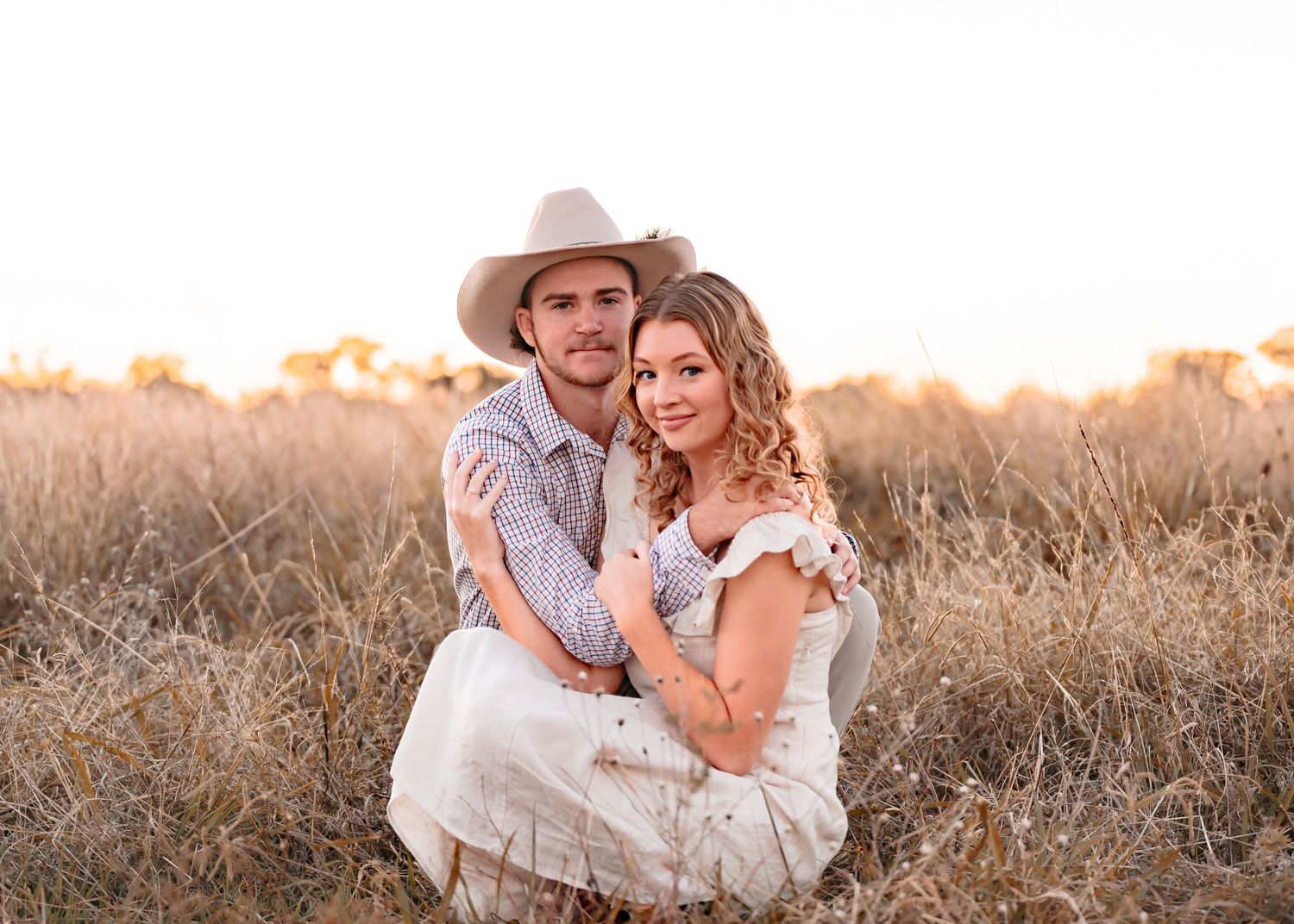 A young, attractive couple kneeling in a vast, open paddock with a beautiful twilight glow. The male has a cowboy hat & is cuddling partner