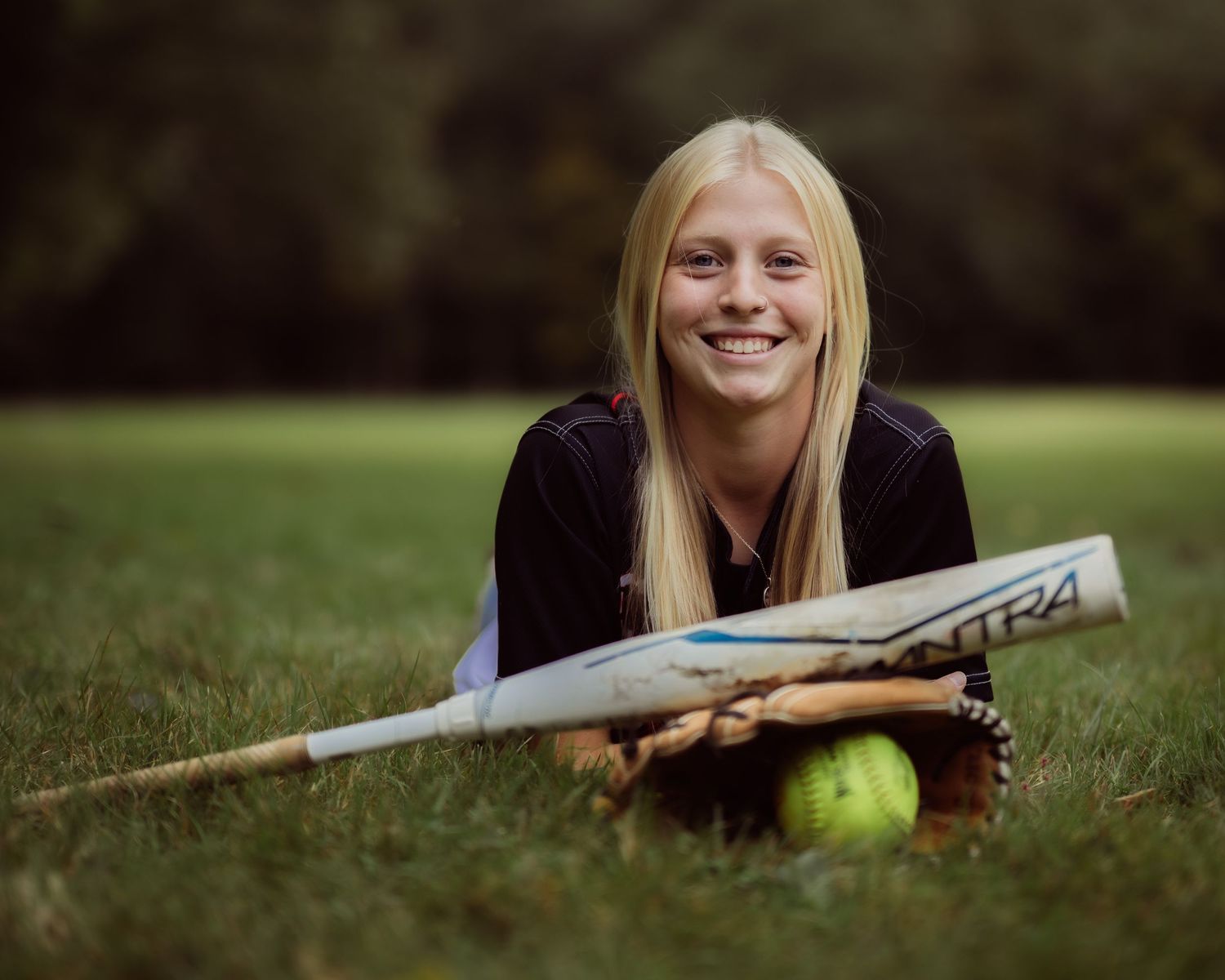 Smiling blonde teenage girl lying on grass with softball bat and glove, posing for sports portrait photo outdoors.
