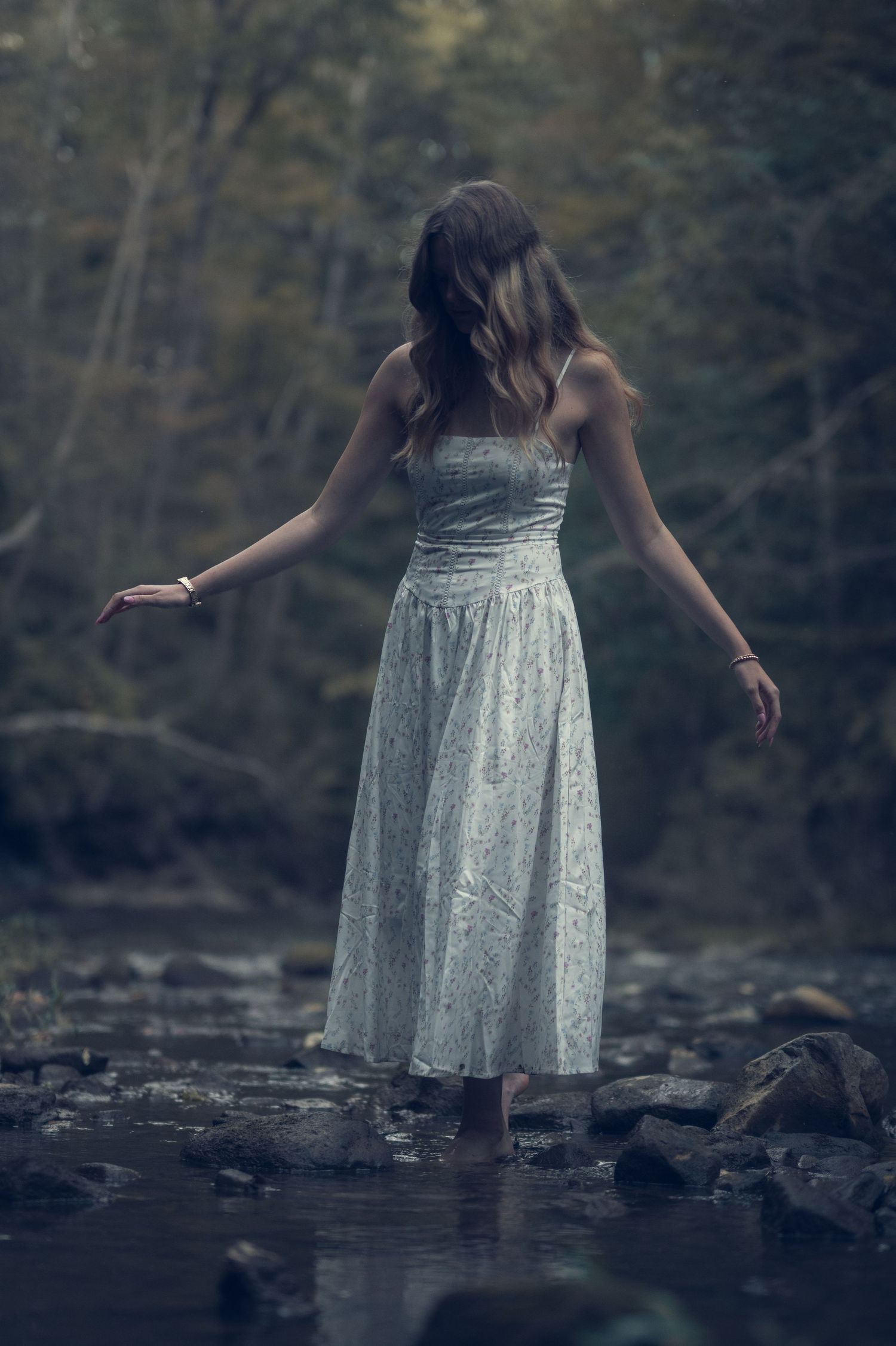 Young woman in white dress standing on rocks in a misty forest stream, arms outstretched.