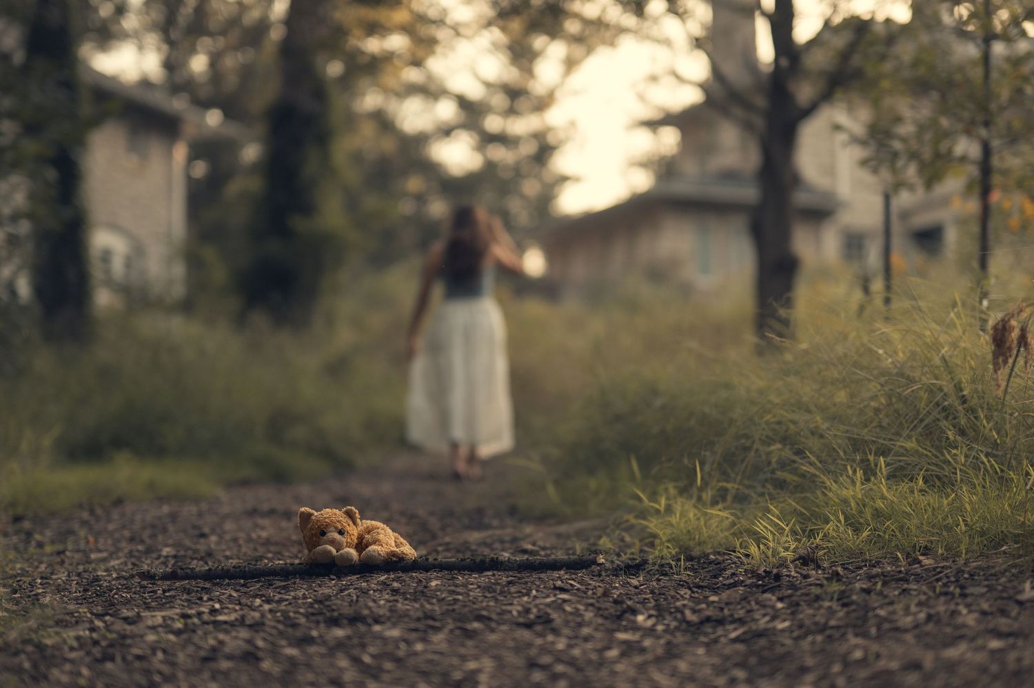 A woman in a white dress walks away on a gravel path, with an abandoned teddy bear in the foreground.