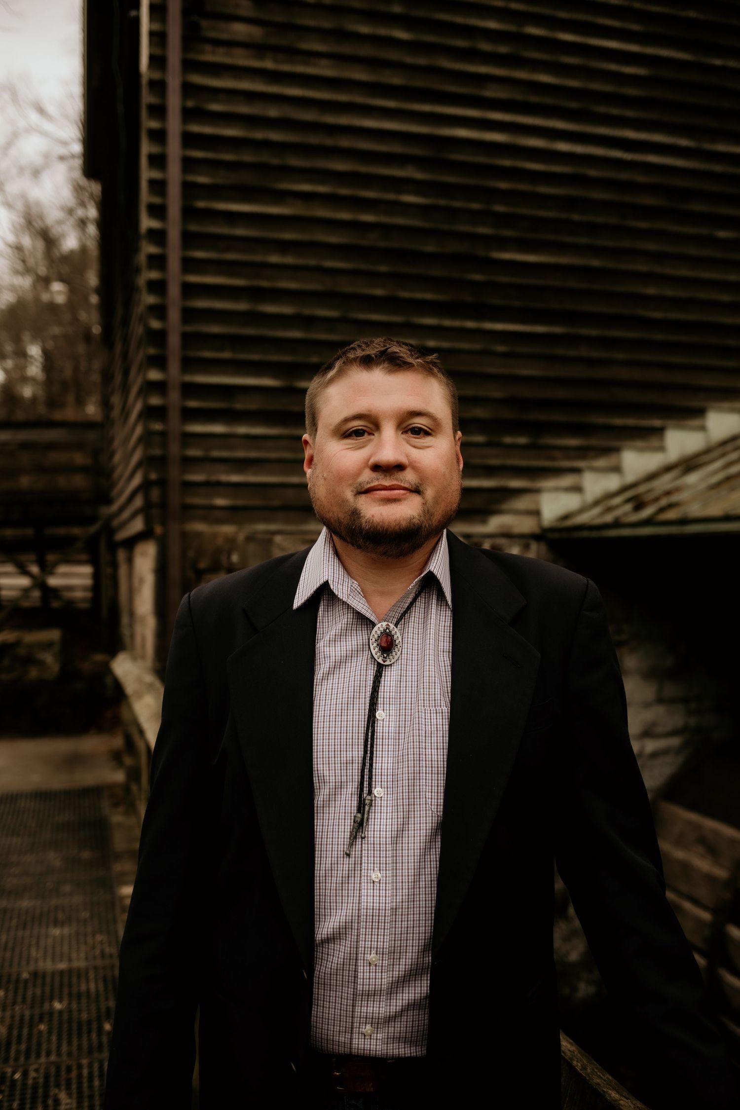 A person in business attire stands in front of a dark wooden building exterior.