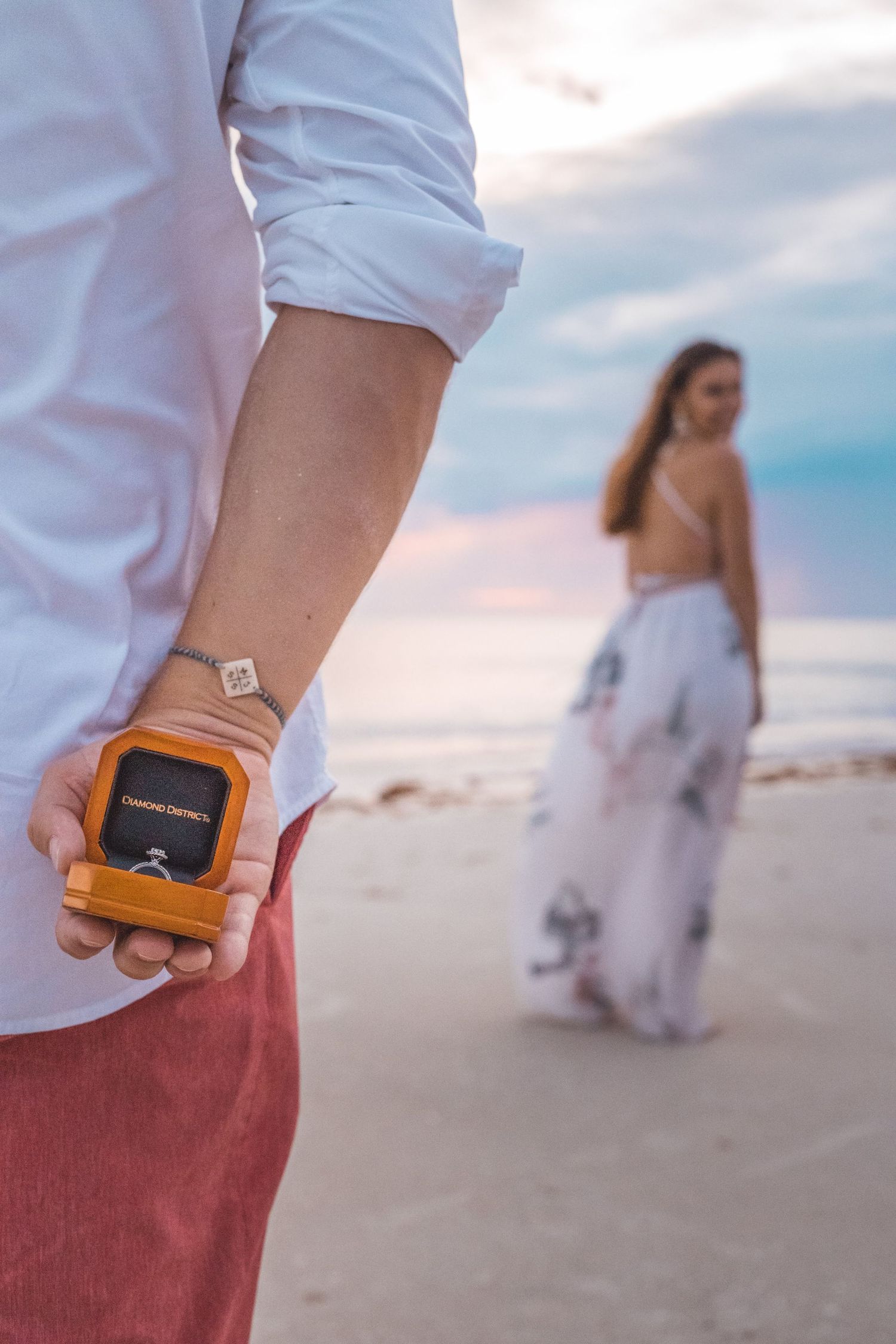 Person holding a small ring box behind their back while standing on a sandy beach.