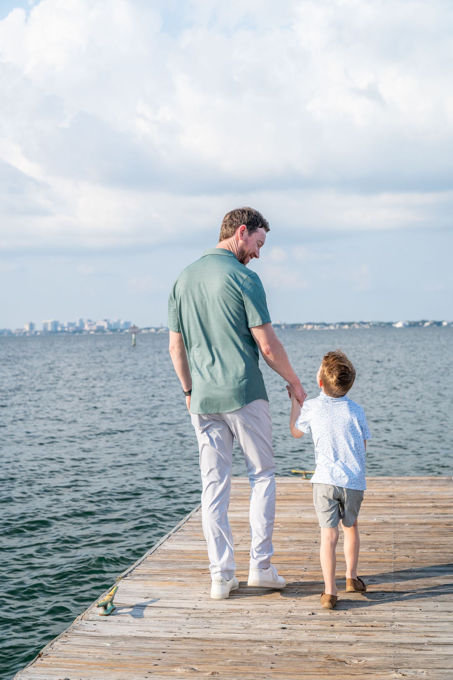 Two people walk hand in hand down a wooden dock overlooking a calm ocean and distant cityscape.