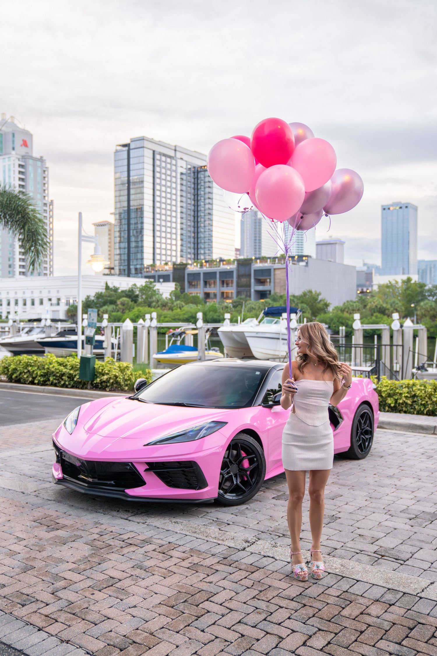 Pink McLaren 720S sports car with pink balloons parked on brick pavement in urban setting with skyscrapers in background.