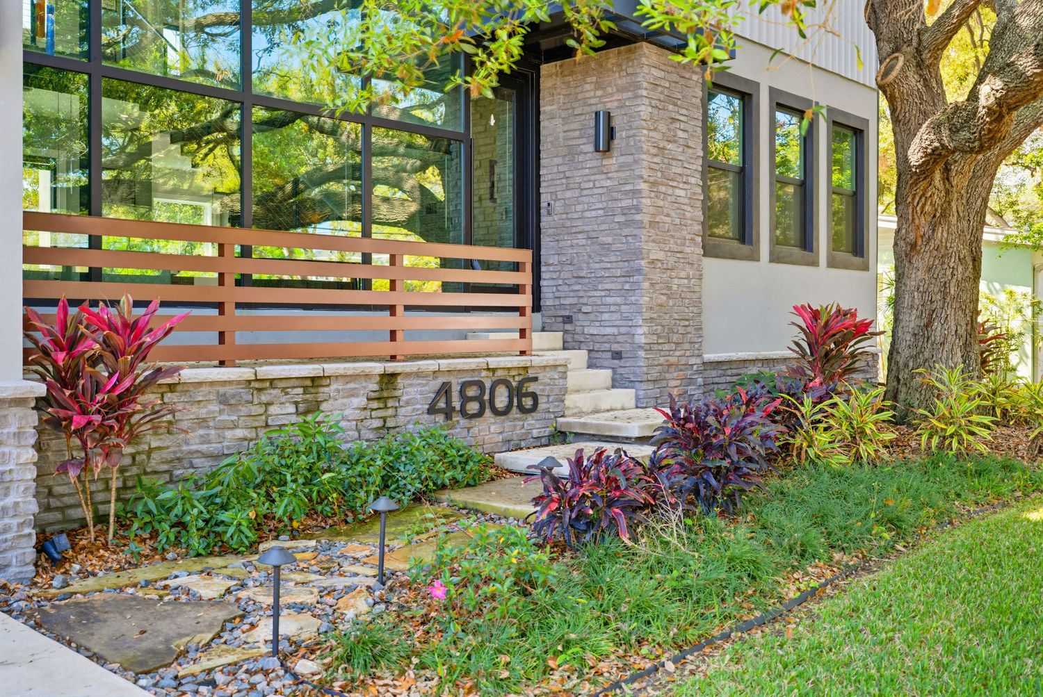 Modern house exterior with stone accents features a well-landscaped front yard with colorful plants and trees.