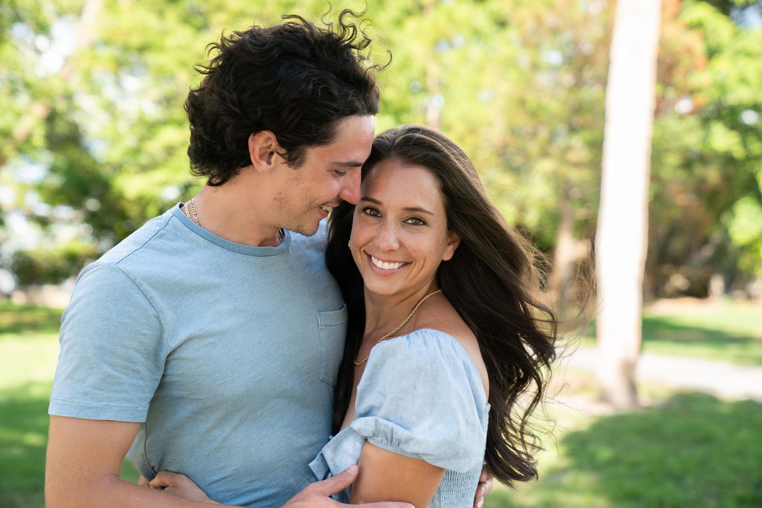 A couple shares tender moments together in a sunny outdoor setting with green trees in the background.