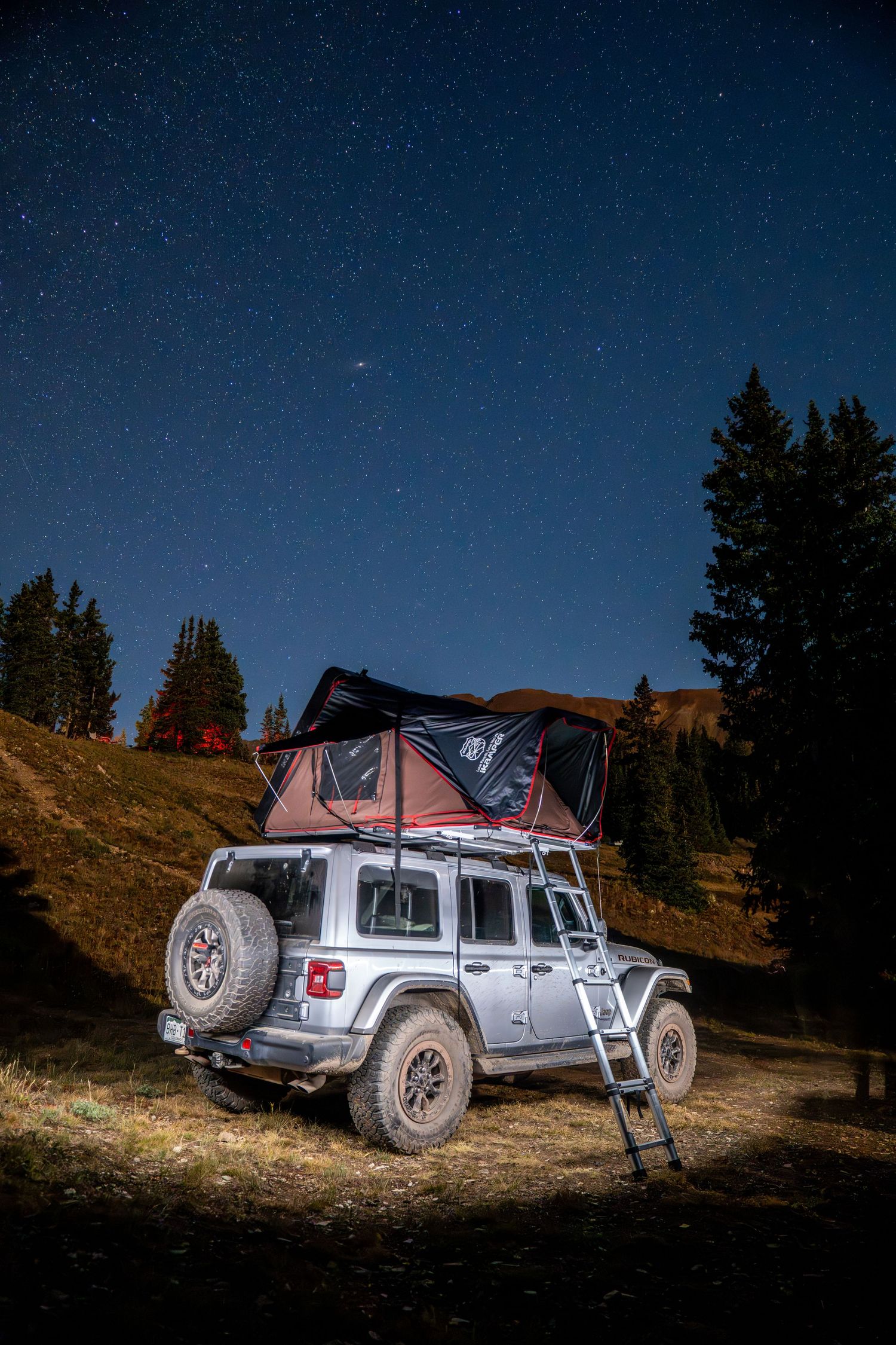 Silver SUV with rooftop tent camping under starry night sky in mountain setting.
