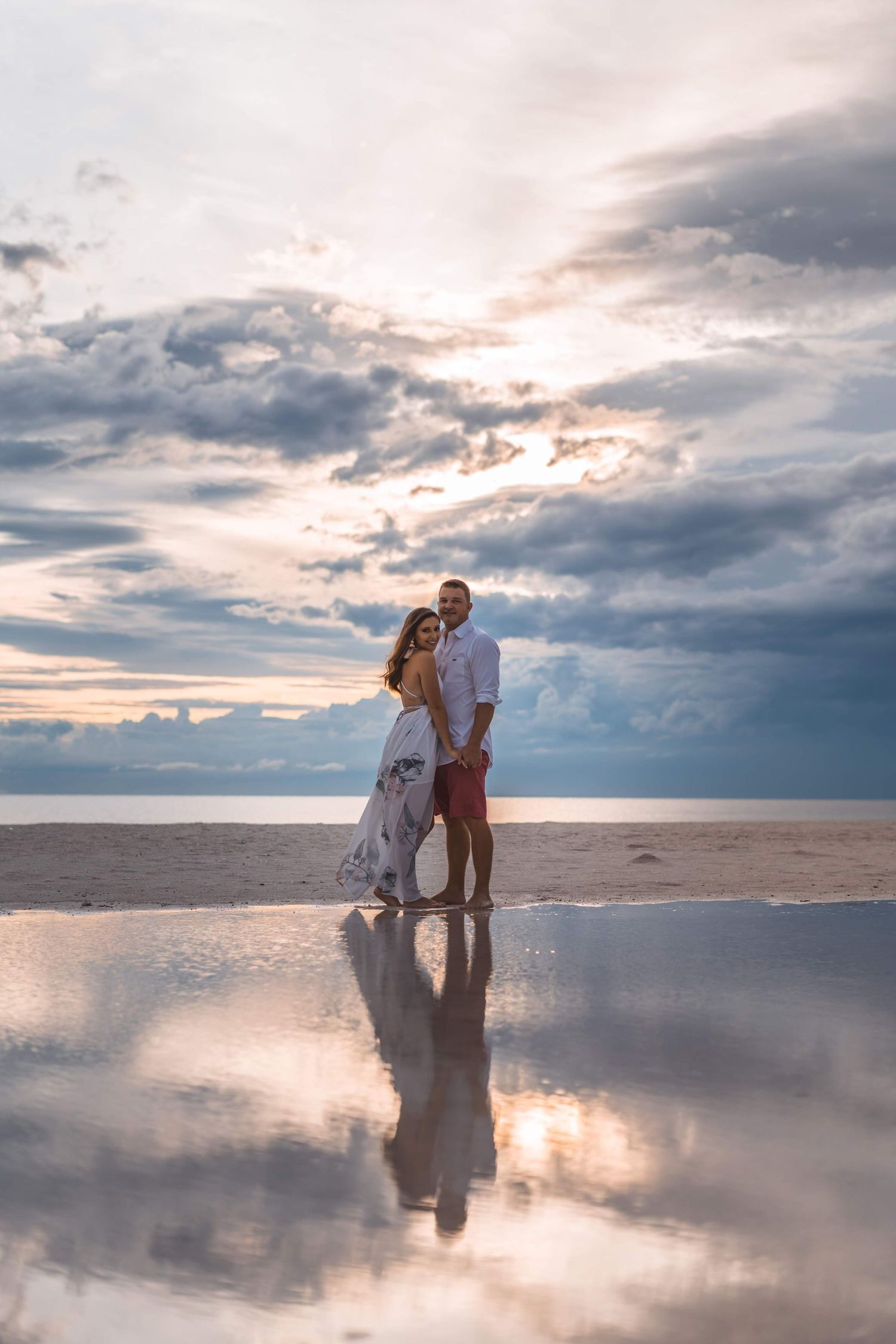A couple embraces on a reflective salt flat during sunset with dramatic cloudy skies overhead.