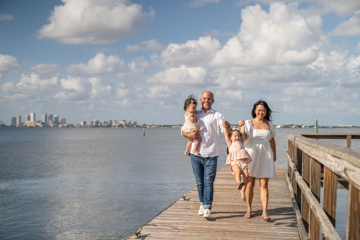Family walking together on a wooden pier overlooking a scenic city skyline on a sunny day.