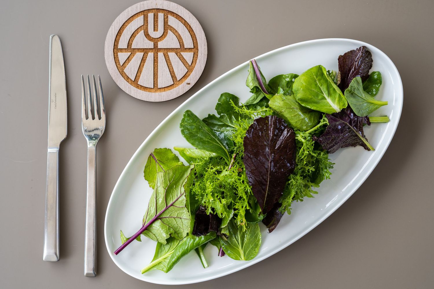 A white oval plate with fresh mixed salad greens and lettuce leaves is presented on a neutral gray surface.