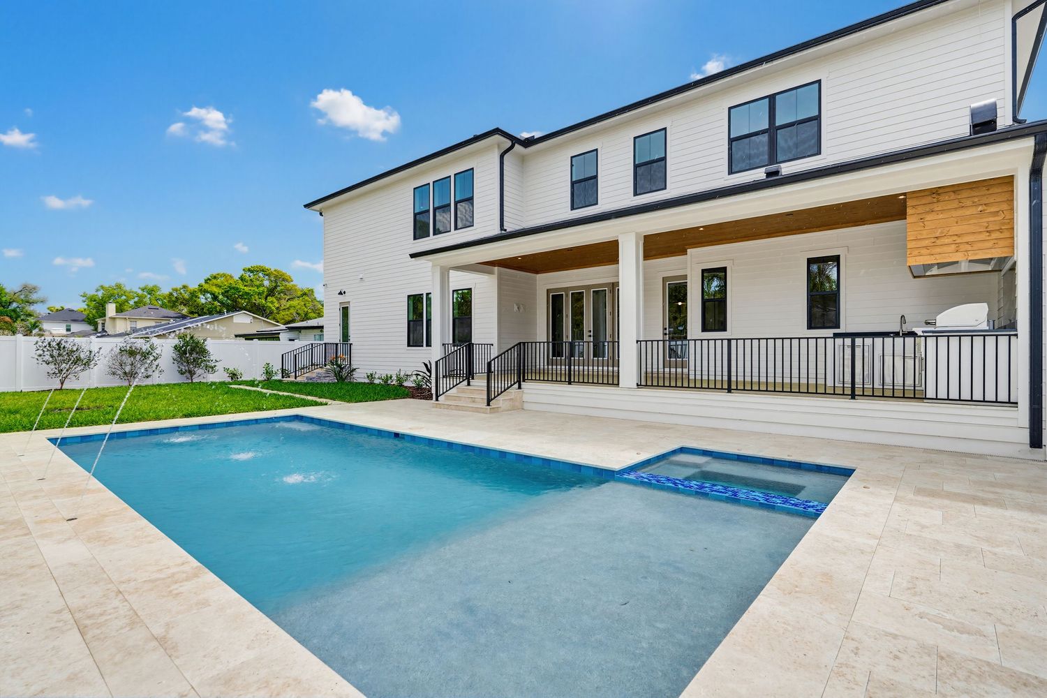 Modern luxury home exterior with swimming pool and travertine deck surrounded by palm trees and blue sky.