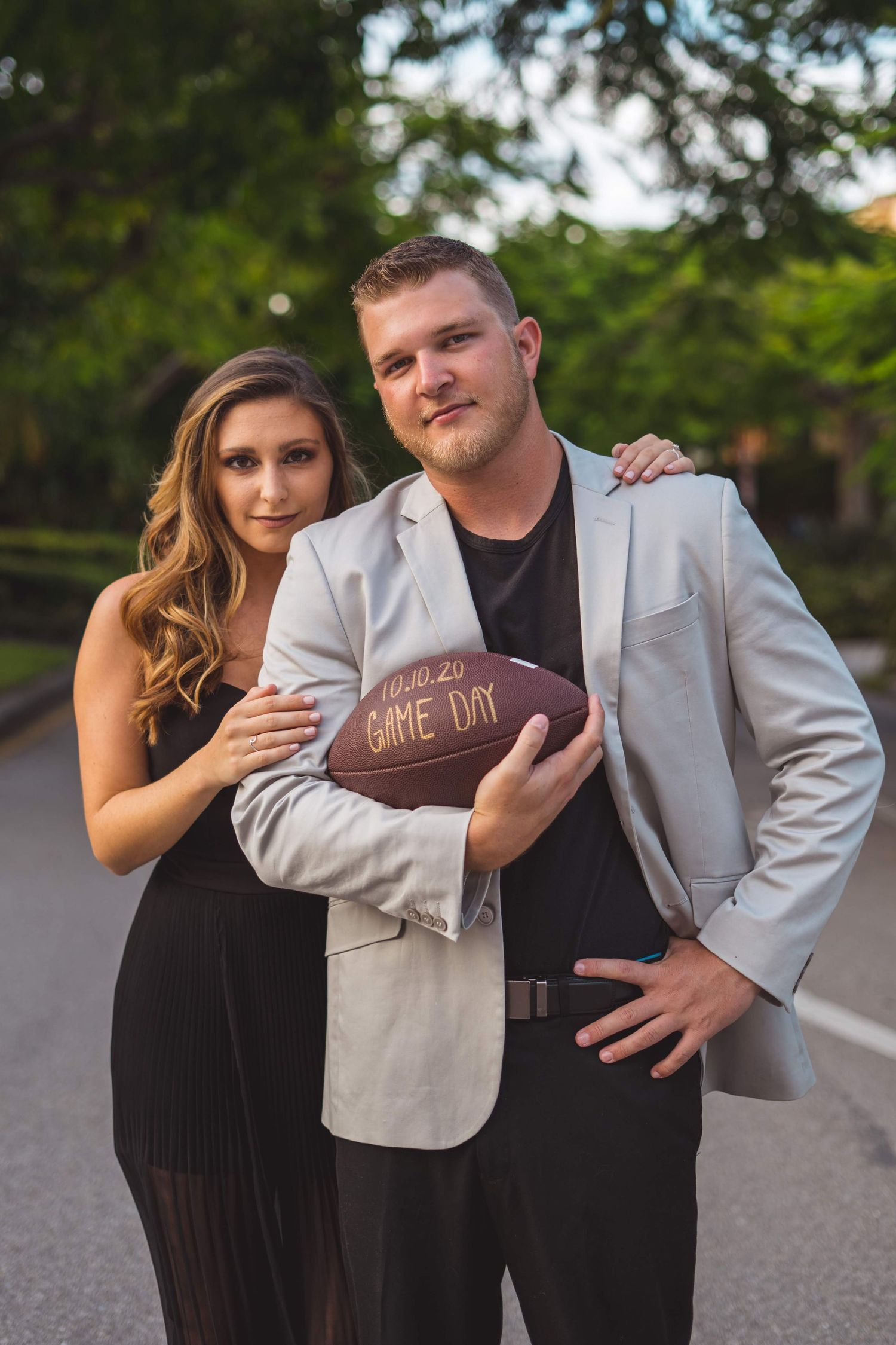 A couple poses together outdoors holding a football in a playful engagement photo shoot.