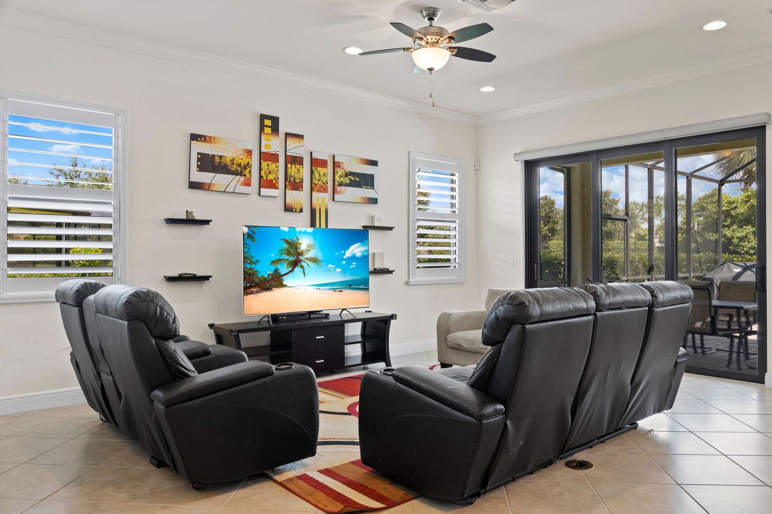 Modern living room with black leather couches, ceiling fan, and large windows overlooking outdoor scenery.