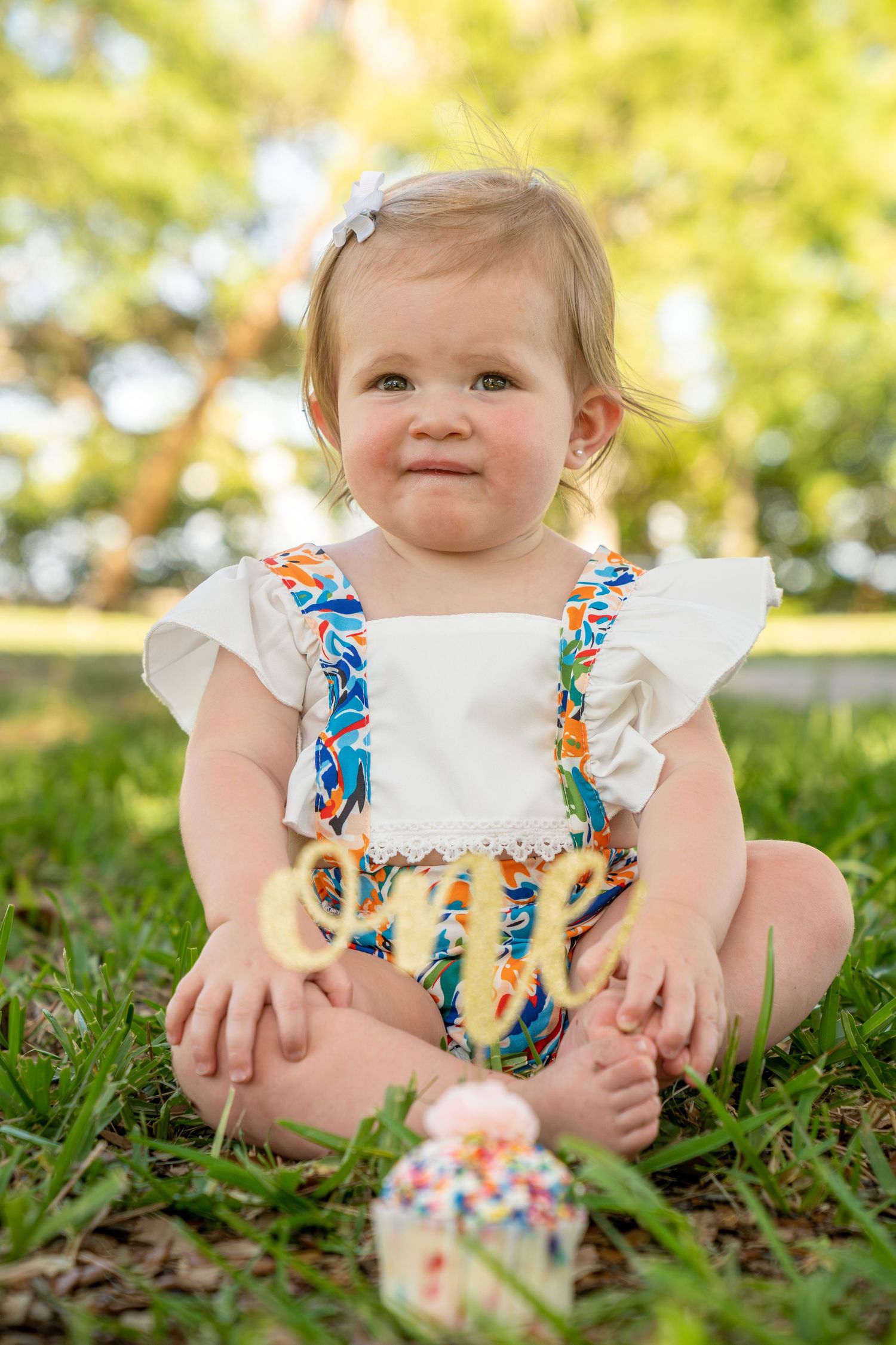 A baby in a white ruffled dress with colorful floral accents sits in green grass on a sunny day.