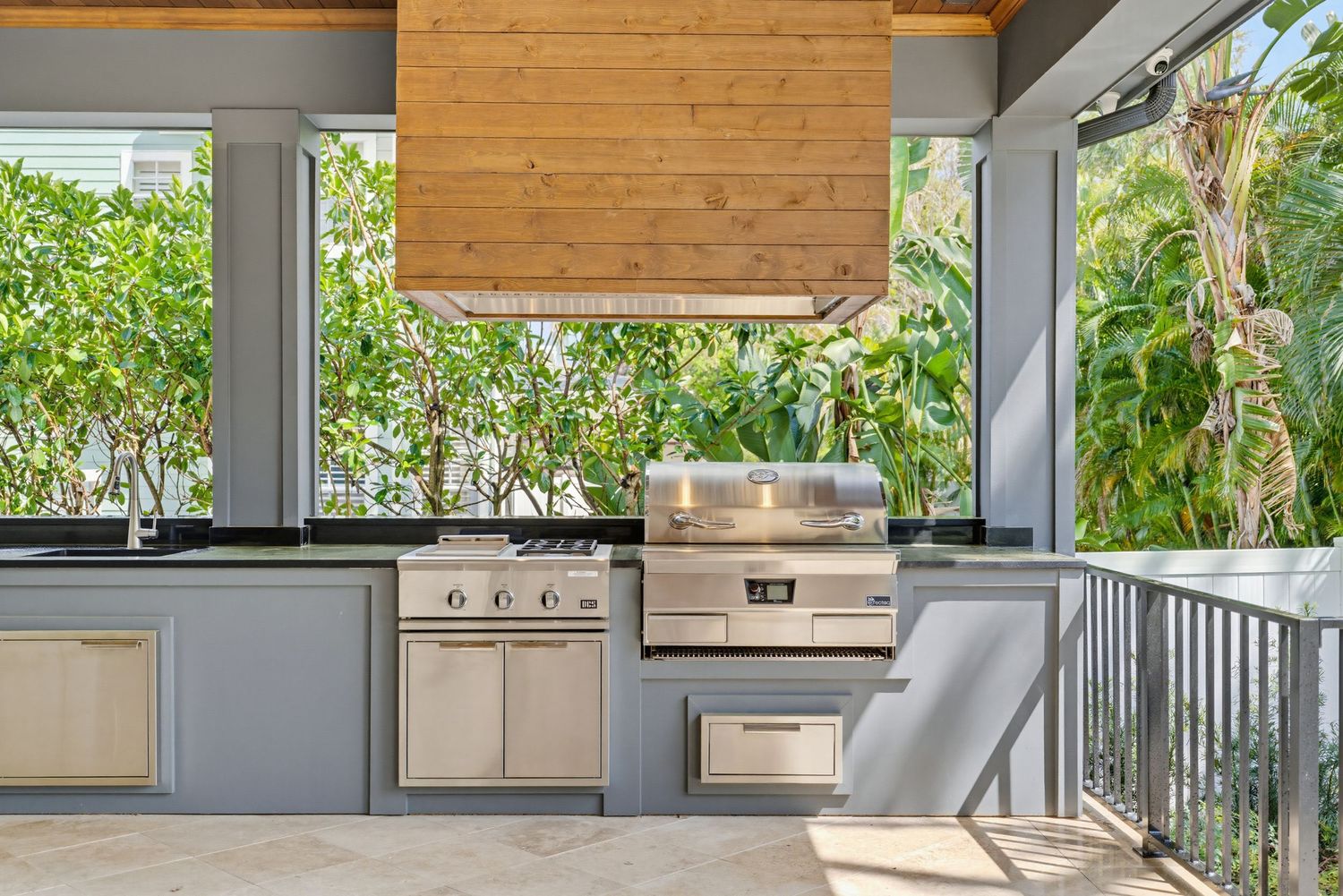 Modern outdoor kitchen with stainless steel appliances and grey cabinets on covered porch surrounded by lush greenery.