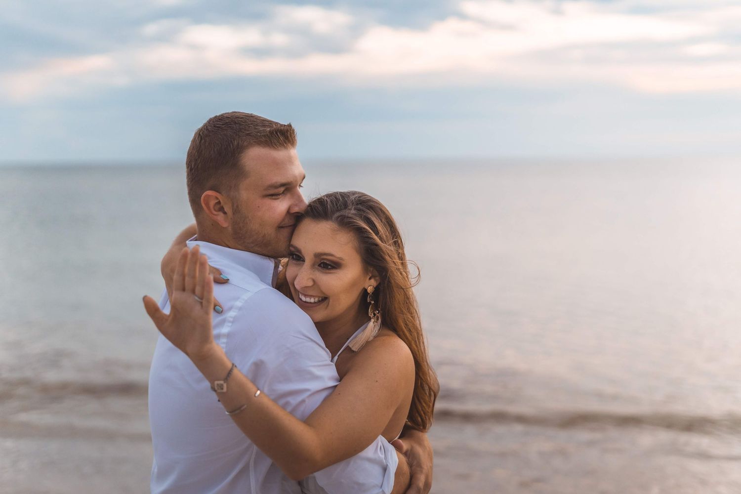 A romantic couple embraces on a sandy beach at sunset with ocean waves in the background.