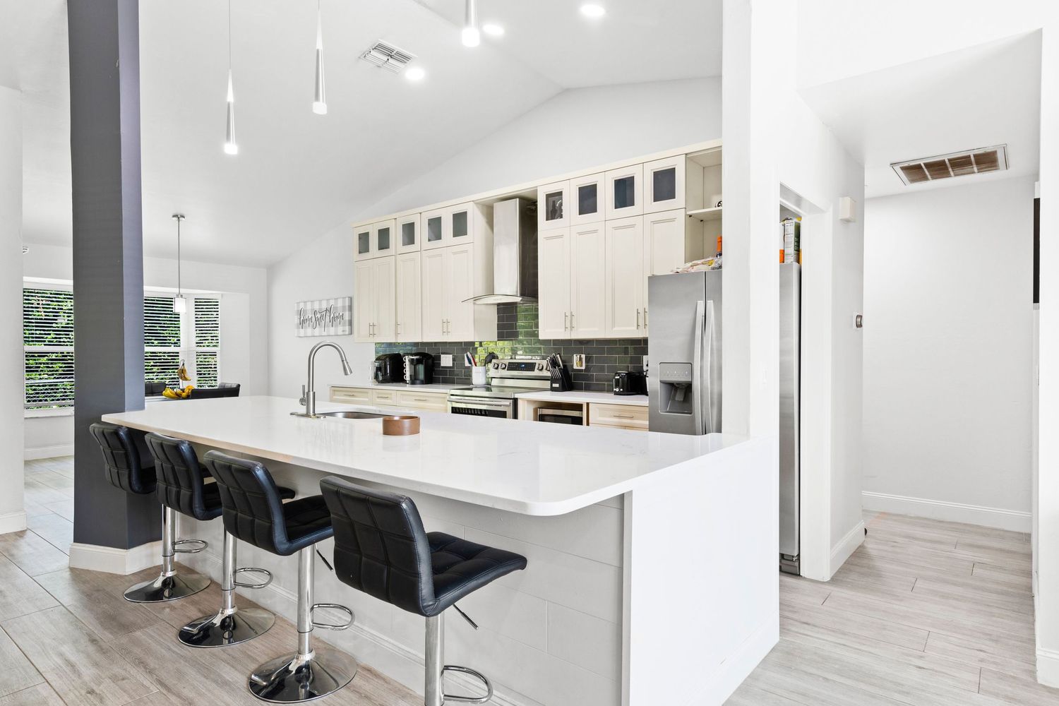 Modern white kitchen with sleek black barstools, quartz countertops, and stainless steel appliances in an open concept space.