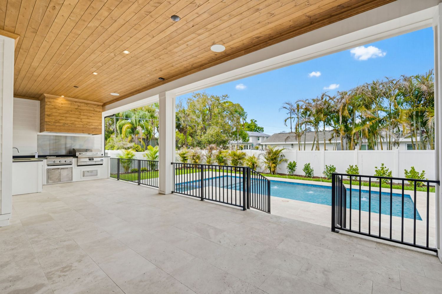 Modern wooden ceiling patio with glass railings overlooks a swimming pool and lush tropical landscaping on a sunny day.