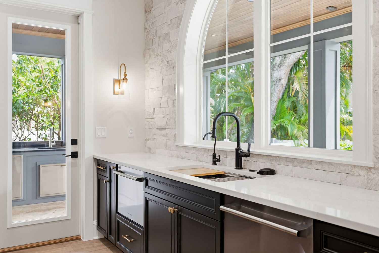 Modern white kitchen with marble countertops, black fixtures, and large windows letting in natural light.