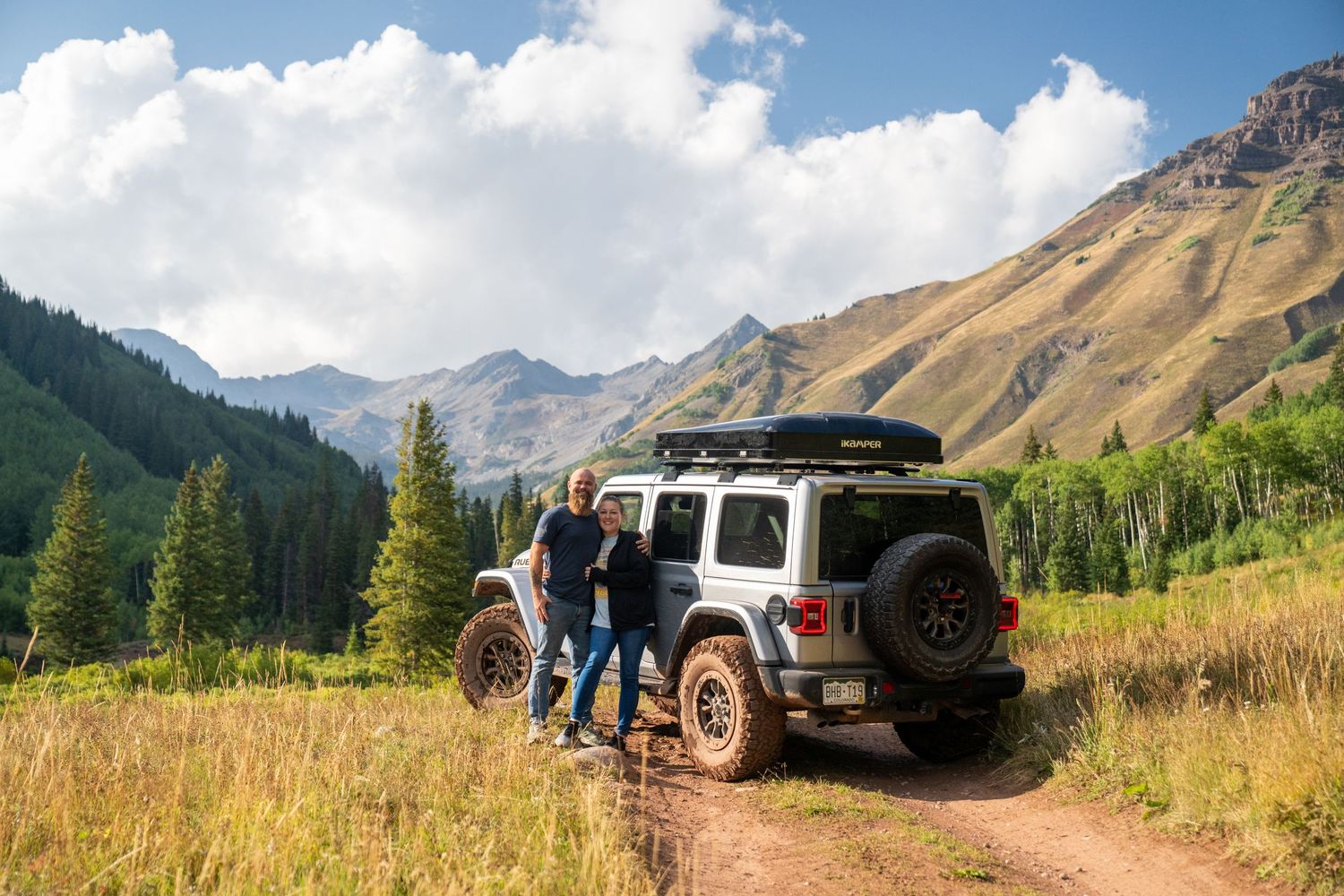 Off-road vehicle parked on mountain trail with dramatic peaks and evergreen forest backdrop.