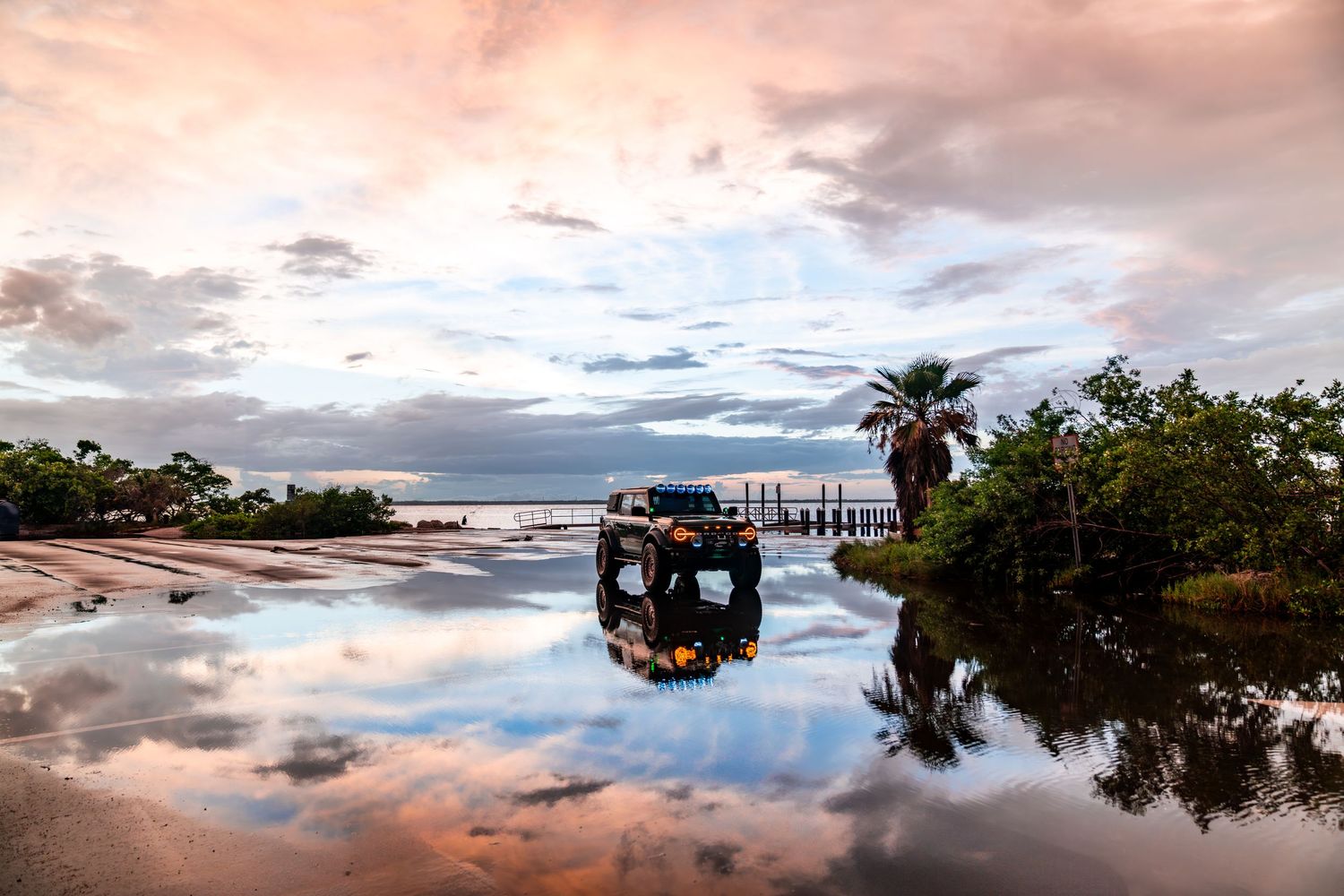 Off-road vehicle creating perfect reflection in coastal waters during sunset with palm trees.