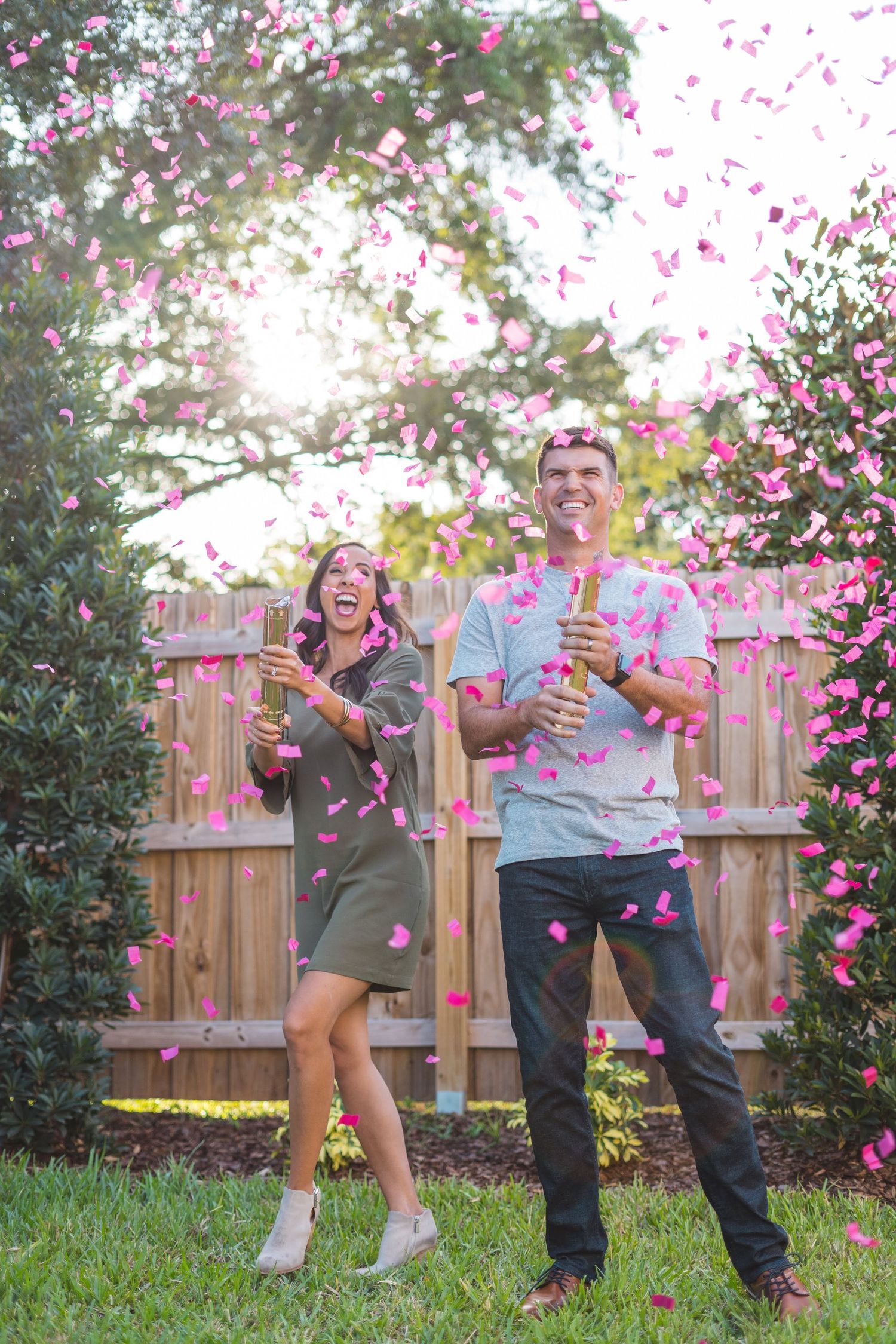 A couple celebrates with pink confetti in a backyard during a special moment by a wooden fence.