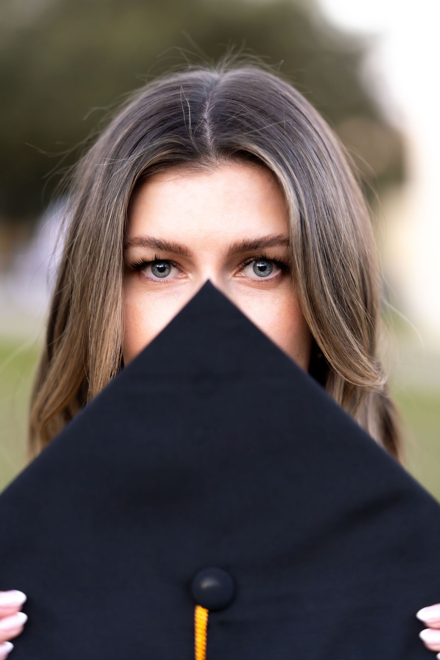A graduation cap held up to cover the lower face, showing bright blue eyes and styled hair above.