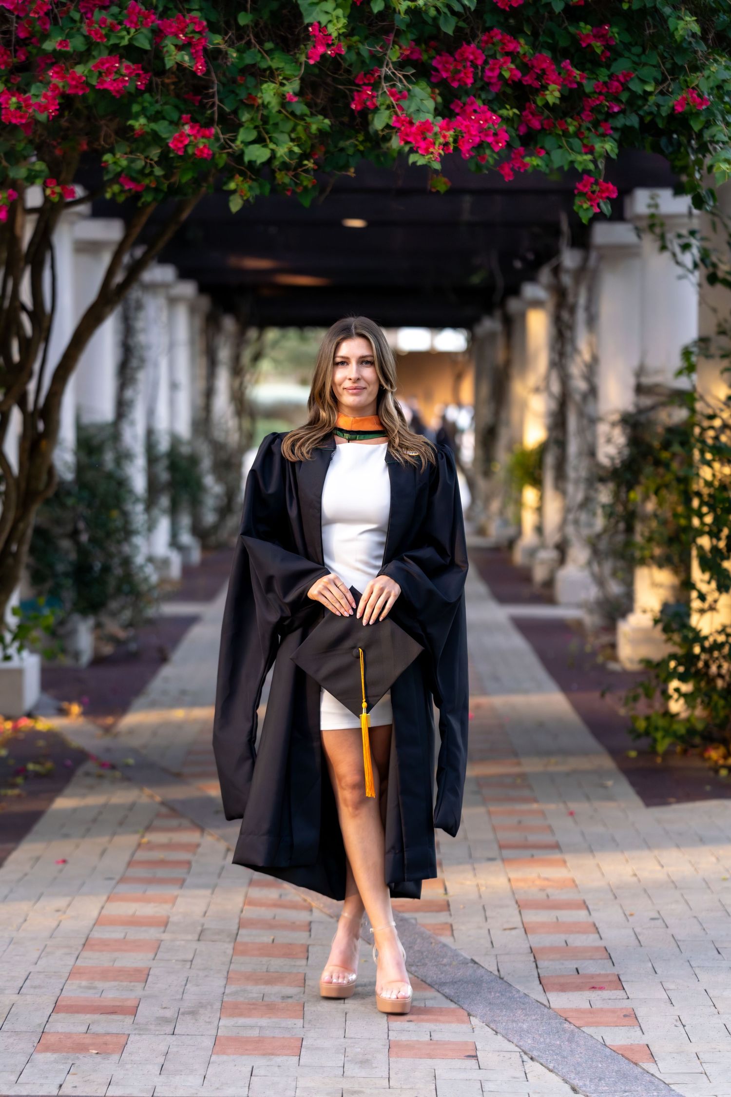 Graduate in black cap and gown poses on brick pathway under flowering archway during graduation photoshoot.