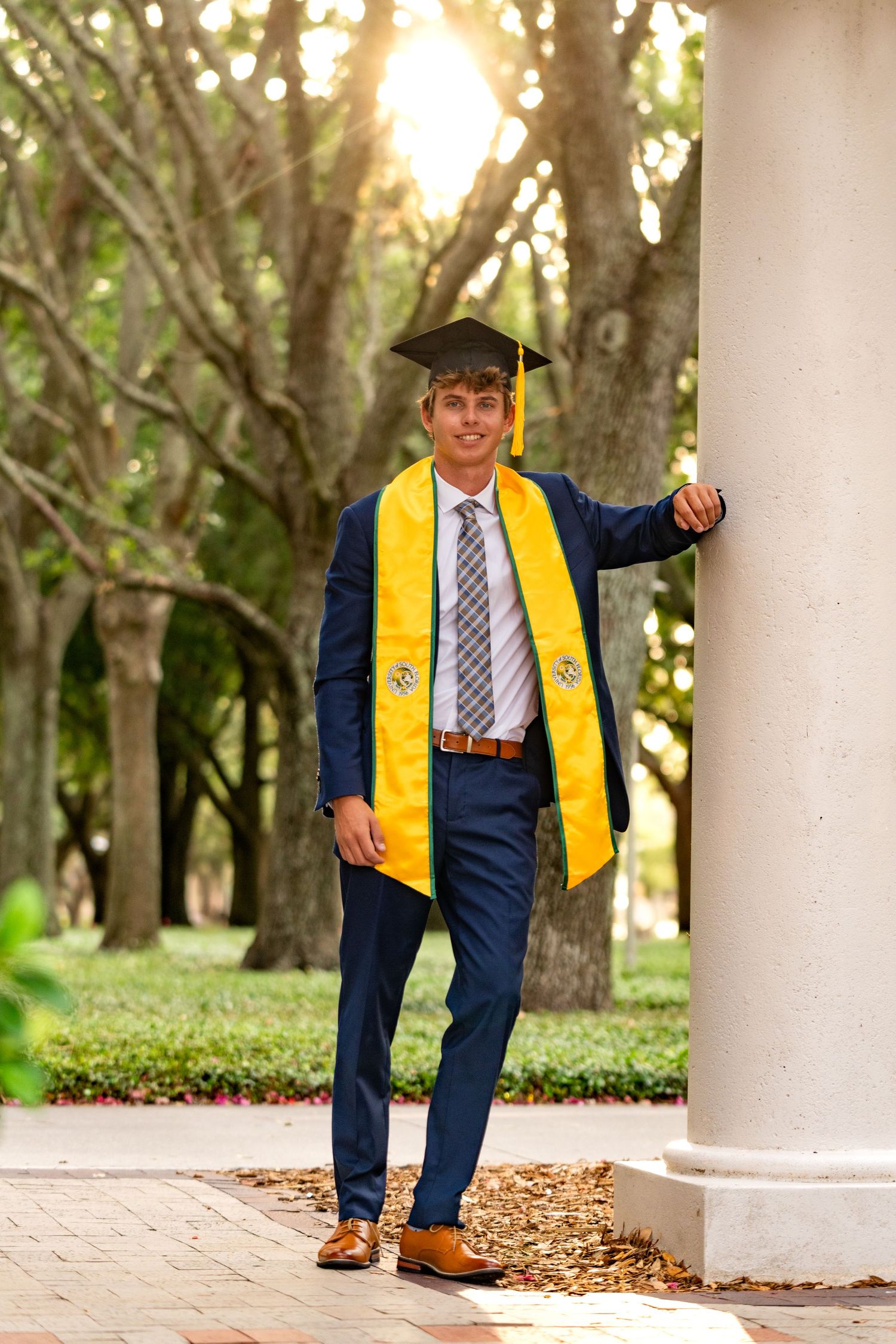 Graduate in navy suit and yellow stole poses playfully behind white column in outdoor garden setting.