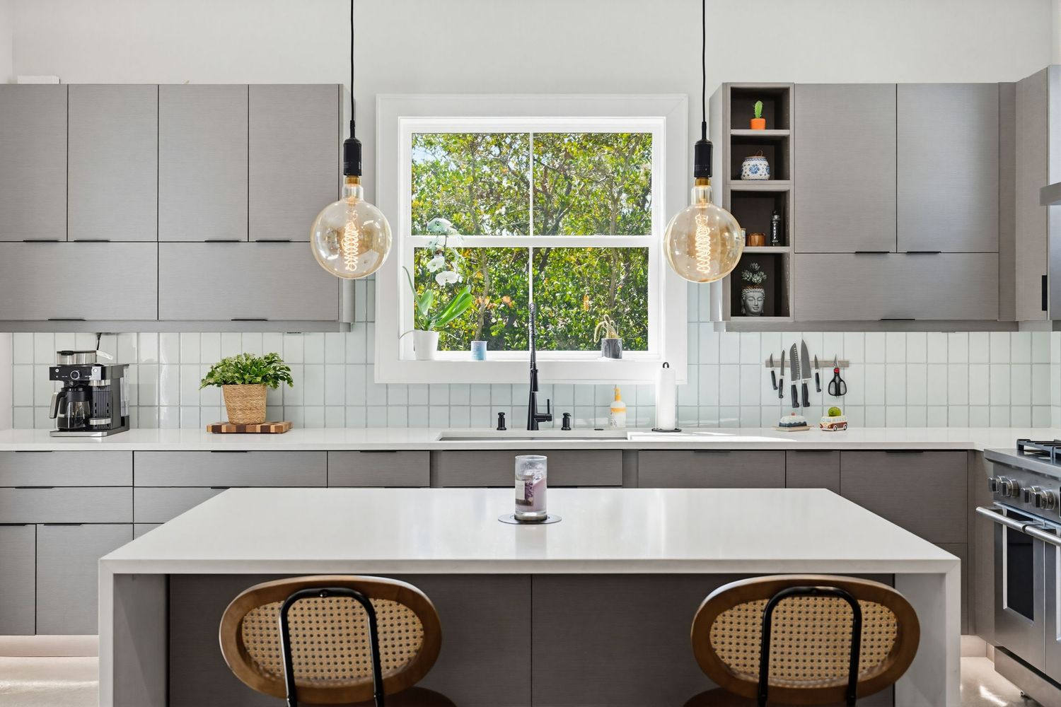 Modern white kitchen with quartz countertops, gray cabinets, and rattan counter stools features natural light from windows.