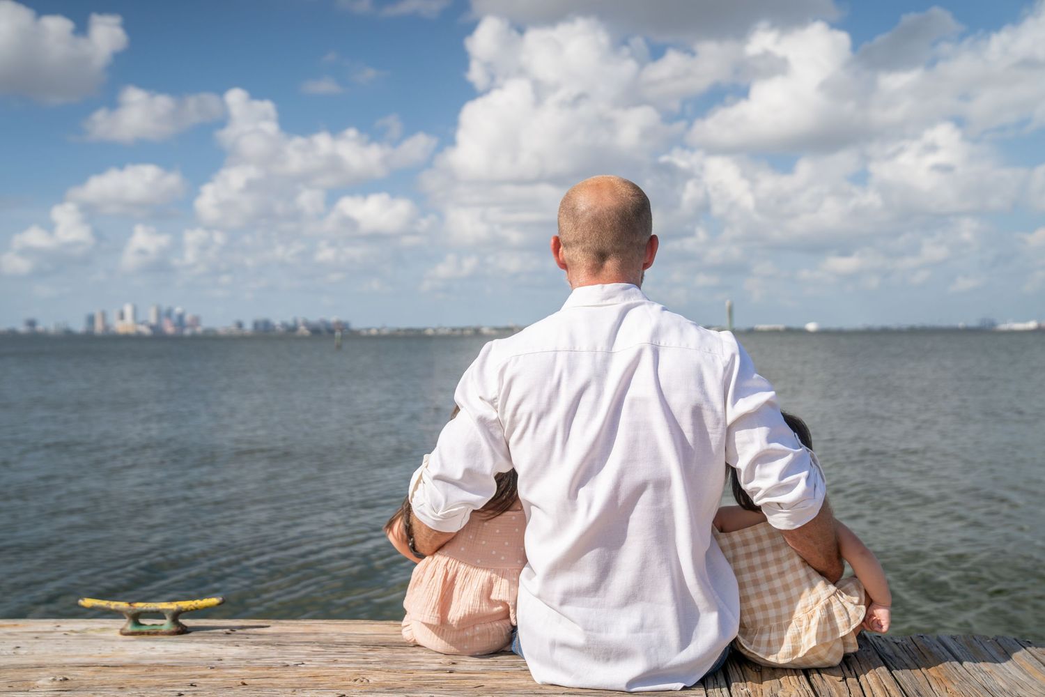 Person in white shirt sits on coastal rocks overlooking calm ocean waters with city skyline visible in distance.